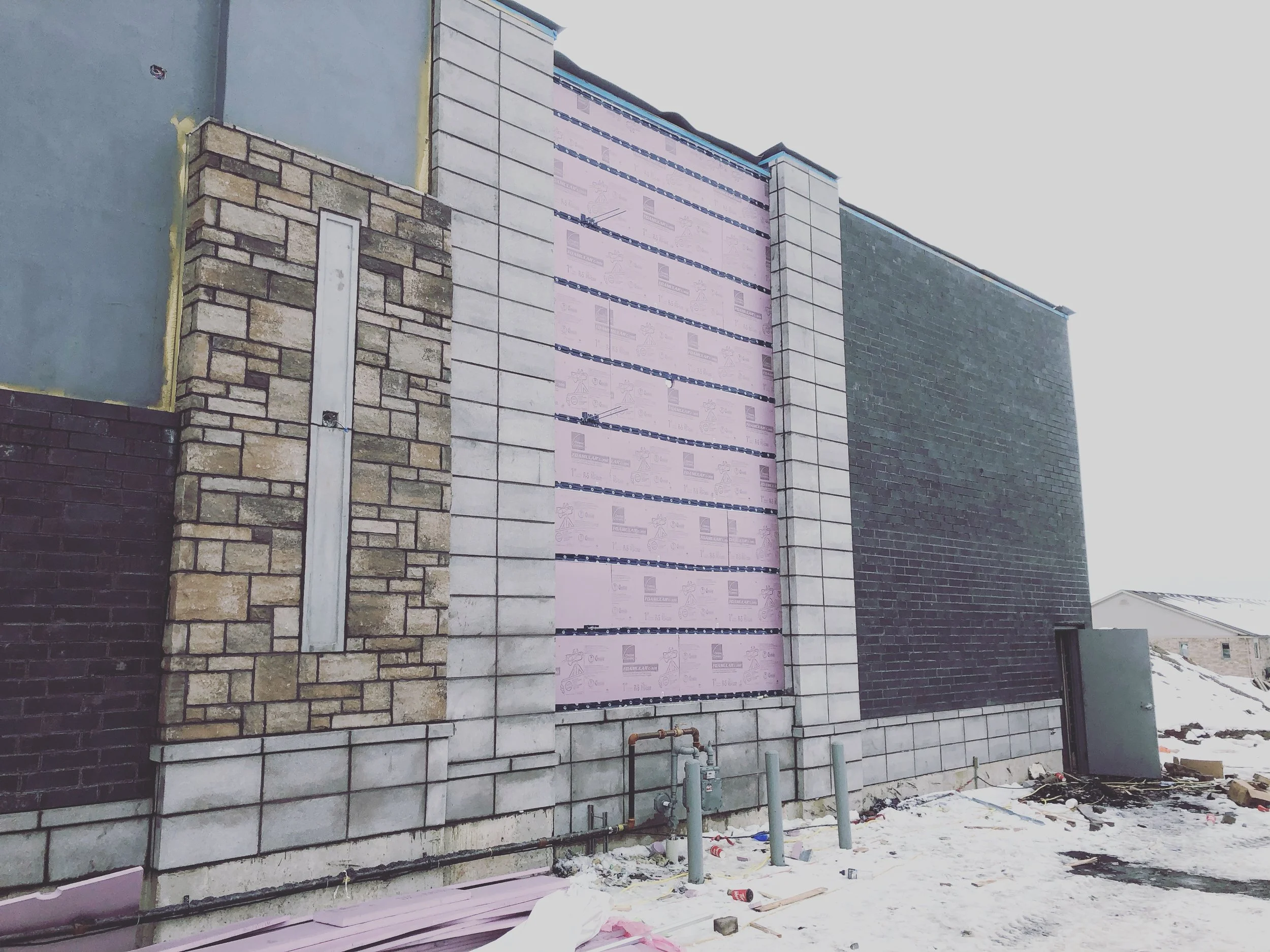 Exterior of a building under construction with different types of wall cladding, including brick, stone, and siding, with construction materials and snow on the ground.