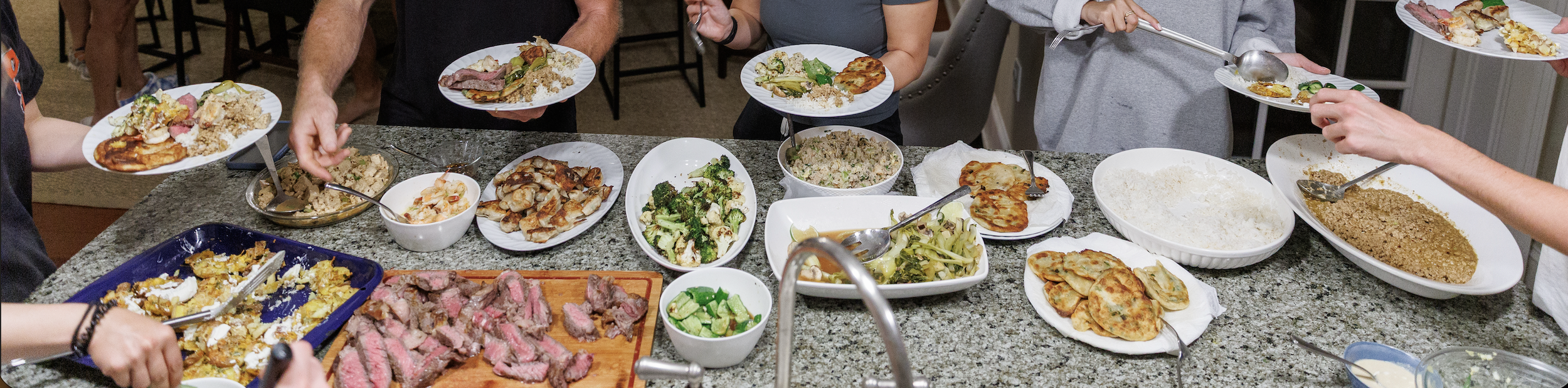 A variety of dishes on a kitchen counter with people serving themselves food