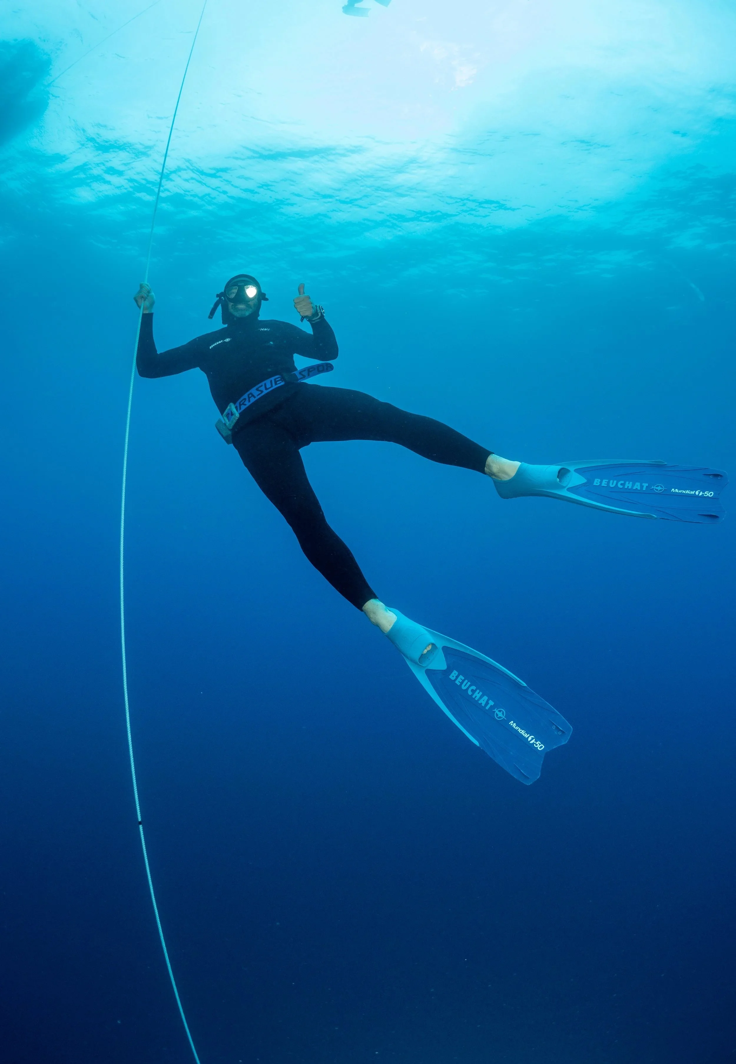 Un plongeur en combinaison noire avec masque et palmes bleues tient une corde en apnée dans l'océan bleu clair, donnant un pouce en signe de positivité.