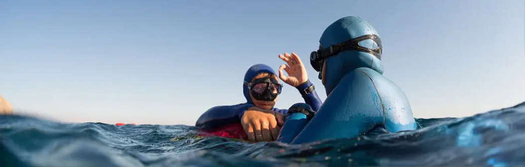 Deux personnes faisant de la plongée sous-marine dans la mer, portant des combinaisons et masques de plongée, en train de communiquer dans l'eau.