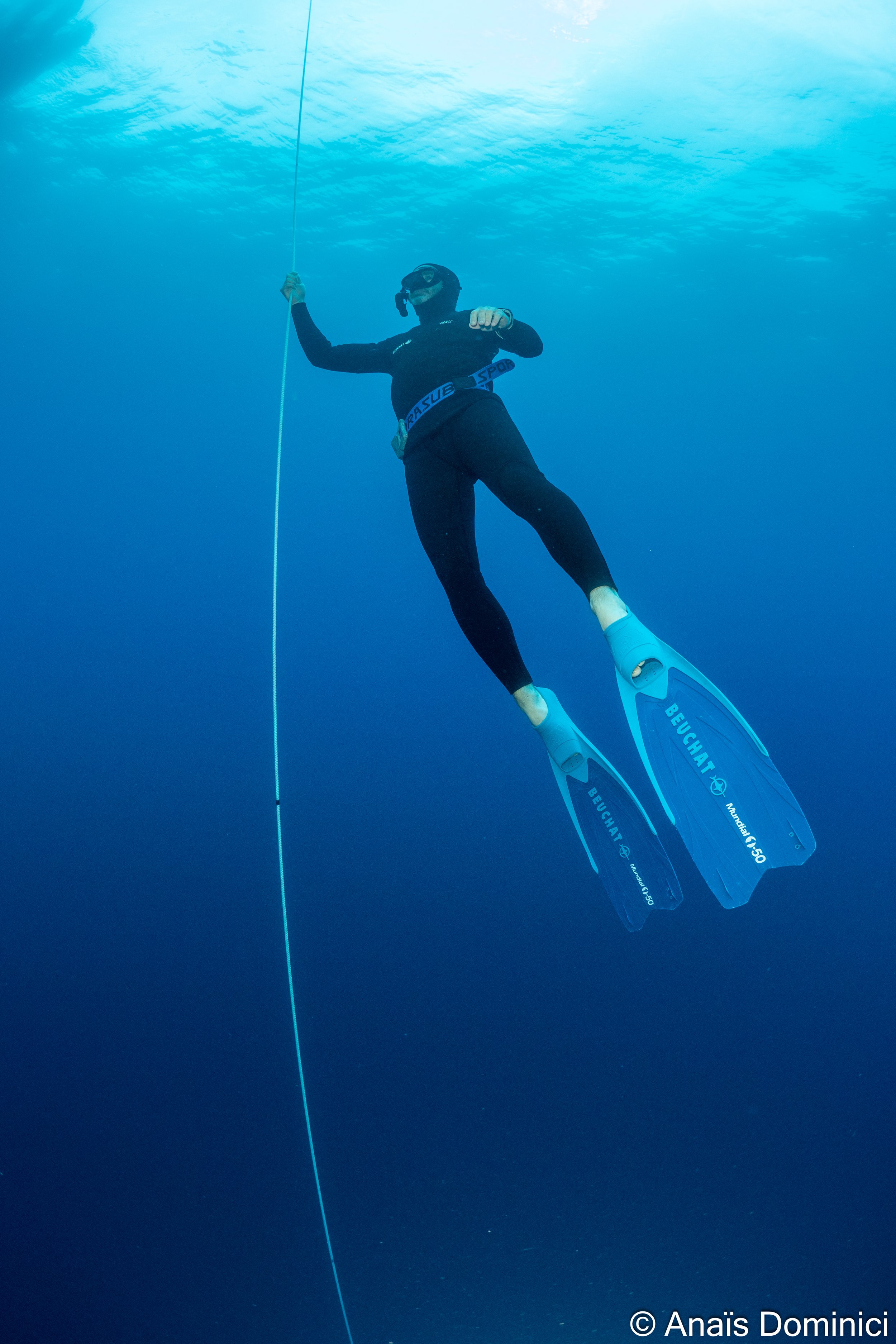 Plongée sous-marine avec un homme en équipement de plongée, nageant en position horizontale, tenant une corde de sécurité, avec des palmes bleues, dans un environnement aquatique clair avec la surface de l'eau visible en haut.