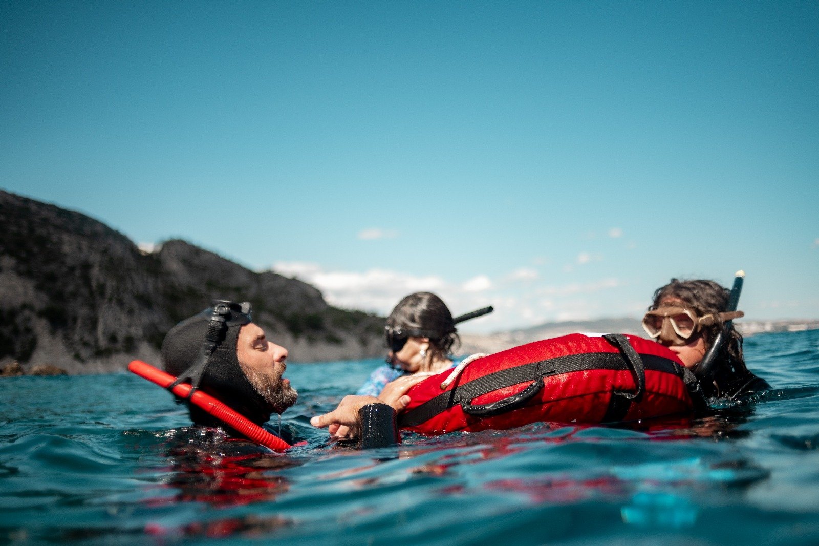 Trois personnes en plongée avec tuba à la surface de l'eau, une portant un masque, une autre un équipement de plongée, avec un paysage rocheux en arrière-plan et un ciel bleu.