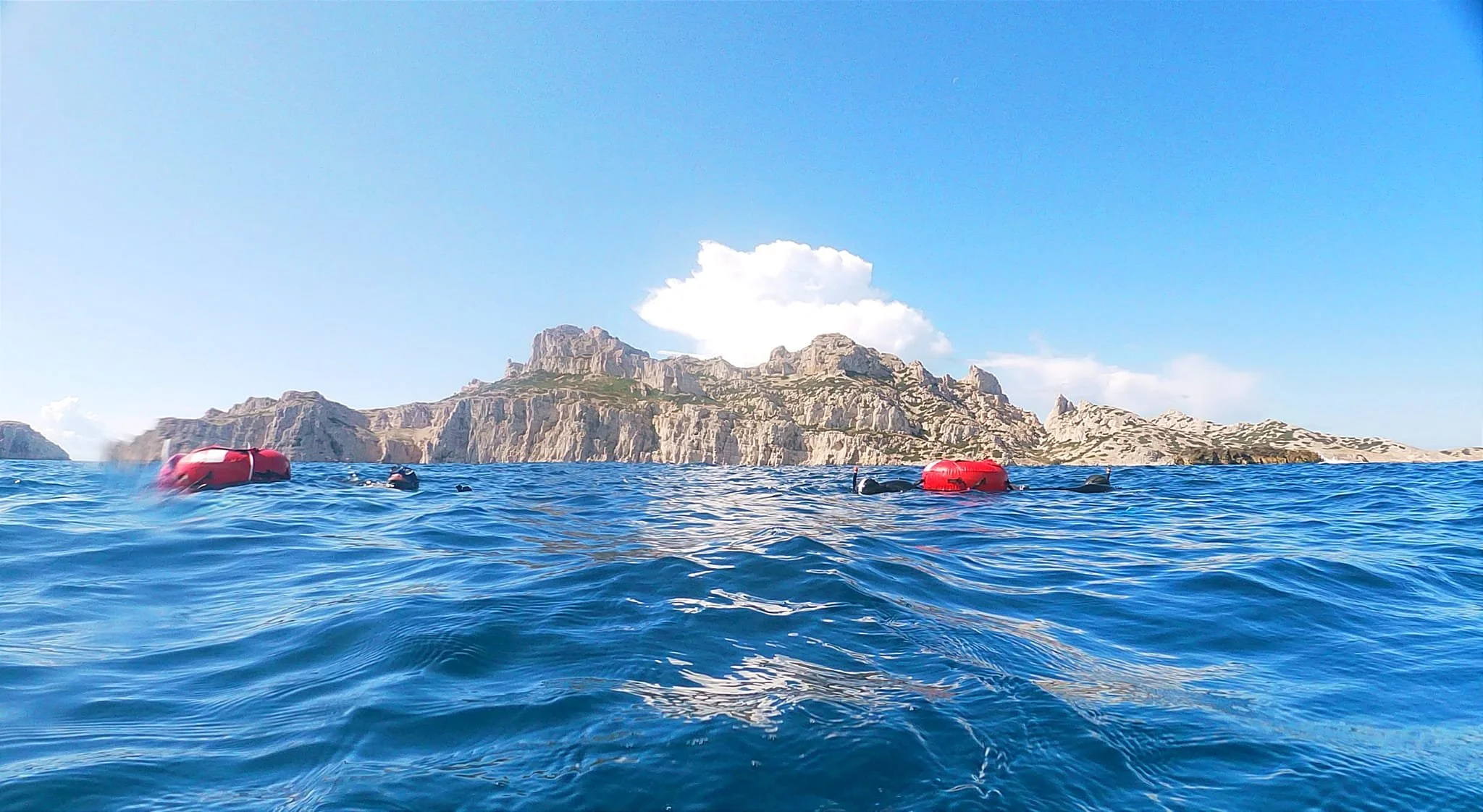 Vue de l'océan avec des bouées rouges flottant à la surface, en arrière-plan les calanques de Marseille, une des falaises rocheuses sous un ciel bleu .