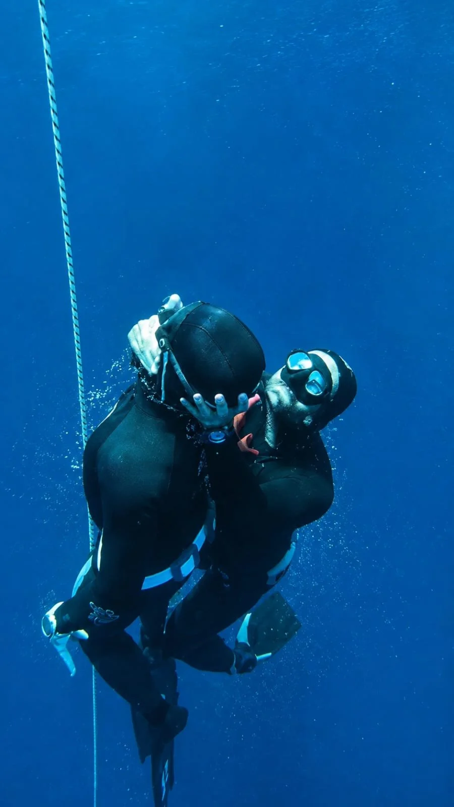 Deux plongeurs en apnée, l'un remonte l'autre en lui bloquant la bouche dans un exercice de safety pour apnéistes au milieu d'un océan bleu profond.