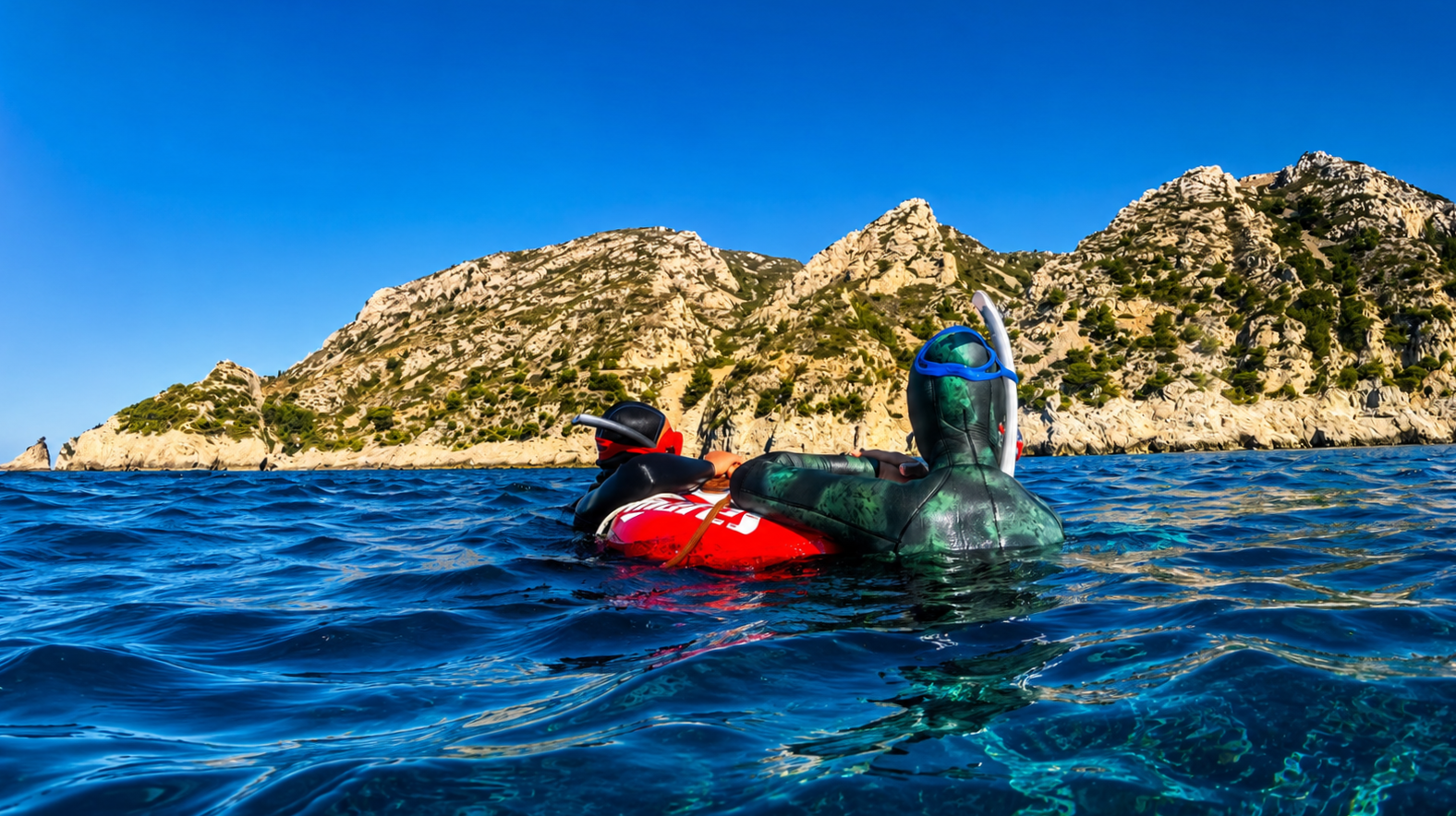 Deux personnes en combinaison de plongée flottant sur la mer, avec une île rocheuse en arrière-plan et un ciel bleu.