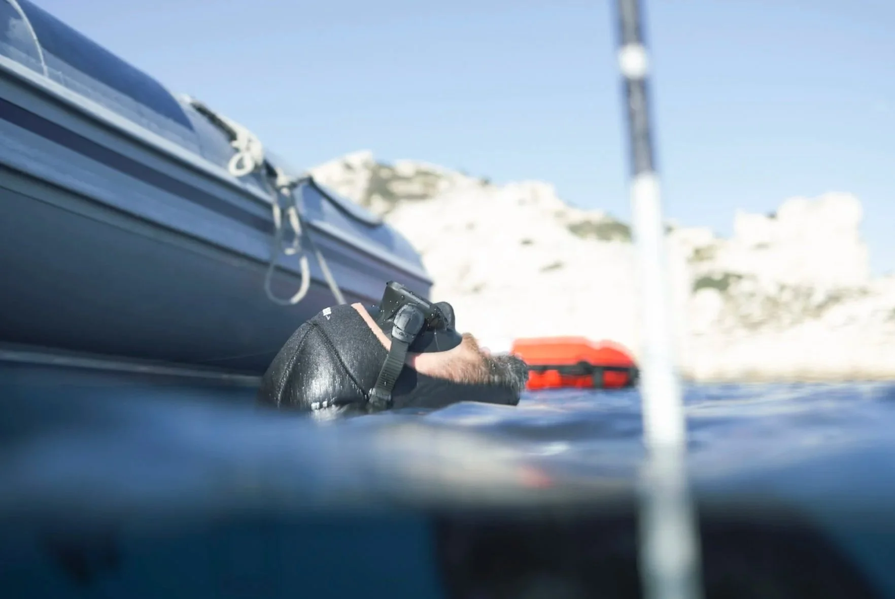 Une main portant un gant noir, manipulant une caméra ou un appareil photo à la surface de l'eau, avec une structure de bateau ou de plateforme à proximité et une toile de fond de falaises ou de formations rocheuses blanches sous un ciel clair.