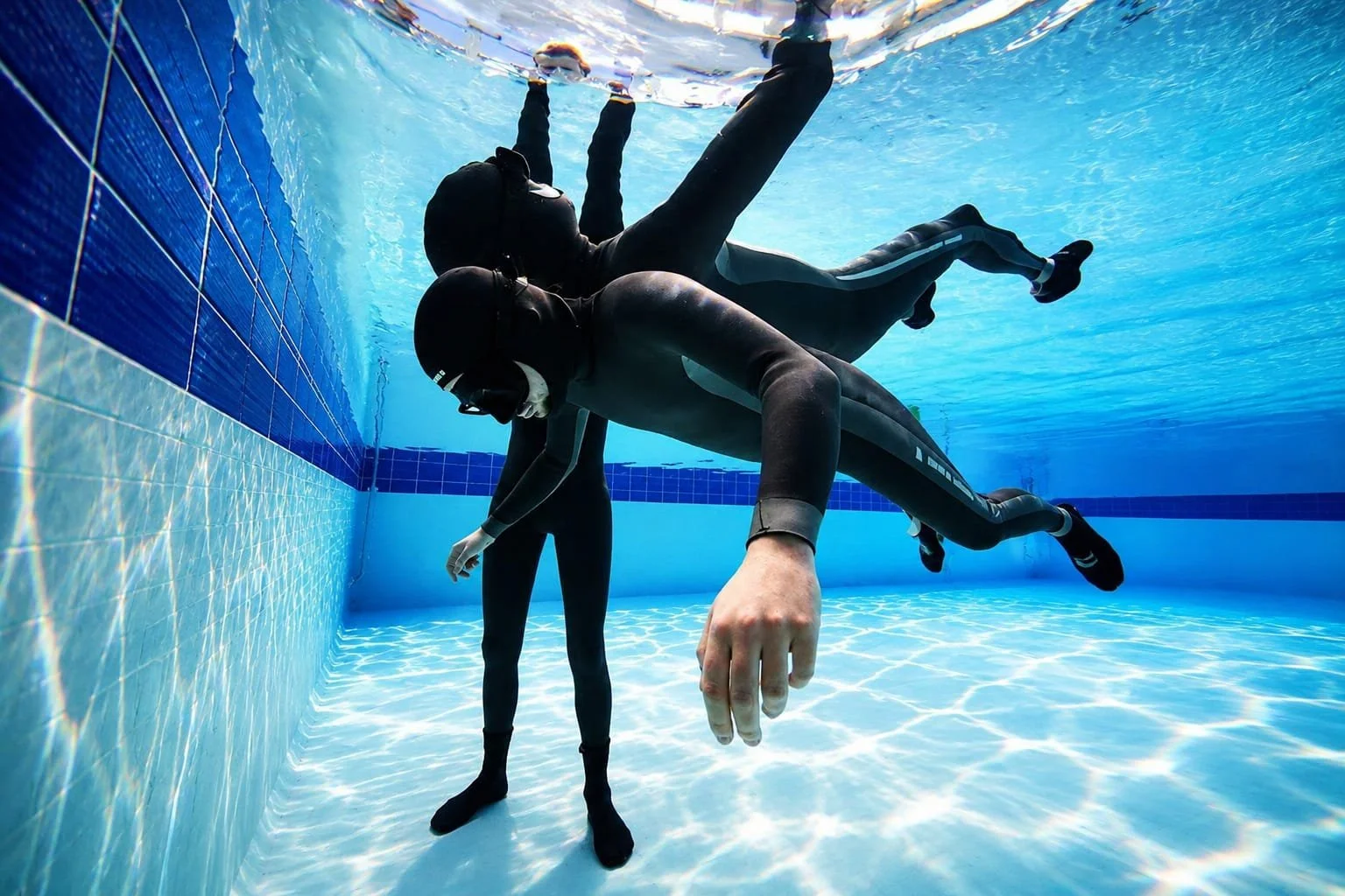 Deux personnes en combinaison de plongée, portant des masques et des tubas, traversent le fond d'une piscine en nageant.