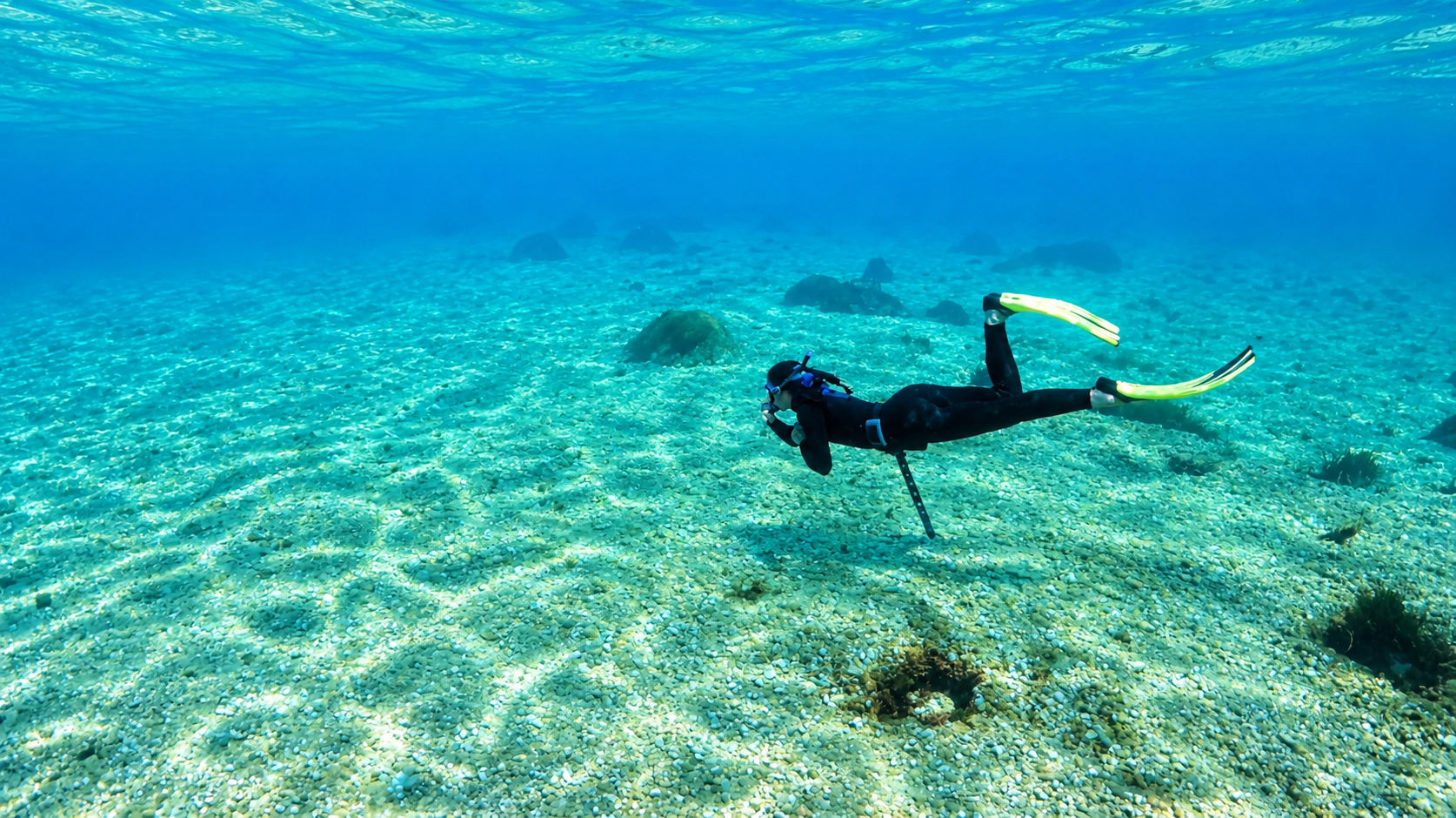 Plongée sous-marine dans une mer claire, avec un plongeur portant un équipement de plongée et palmes, nageant au-dessus d’un fond de sable parsemé de rochers et de quelques végétations