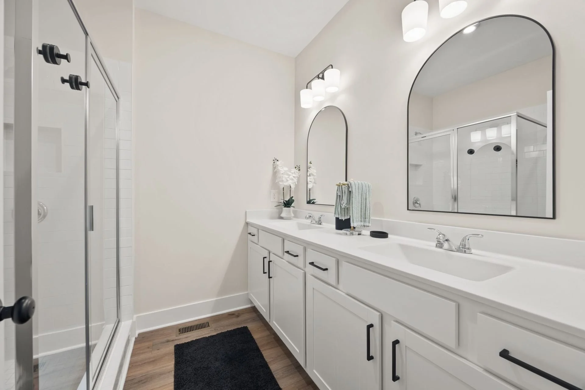 Modern bathroom with double vanity sinks, large mirrors, white cabinets with black handles, overhead lighting, a plant on the countertop, a towel, and a shower with a glass door.