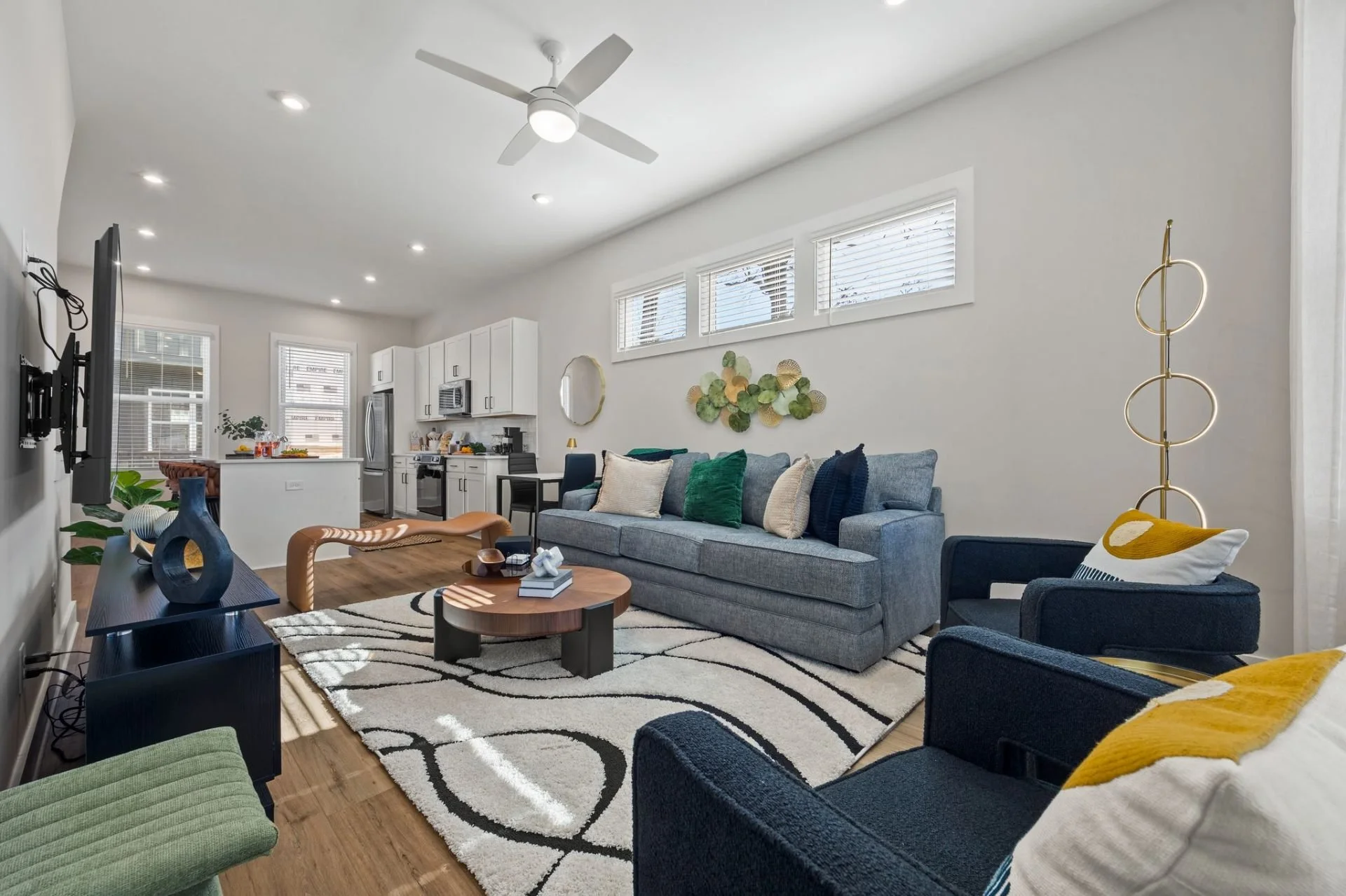 Modern living room with a gray sofa, colorful pillows, a black armchair, and a wooden coffee table on a patterned white and black rug. Open kitchen with white cabinets and stainless steel appliances is visible in the background. Large windows with horizontal blinds let in natural light. Decorative wall art and a ceiling fan are present.