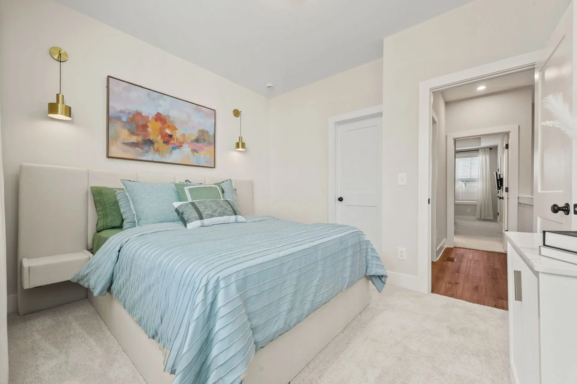 Modern bedroom with a bed featuring blue and green pillows, artwork above the headboard, gold wall sconces, and a view into a hallway with a window and white curtains.