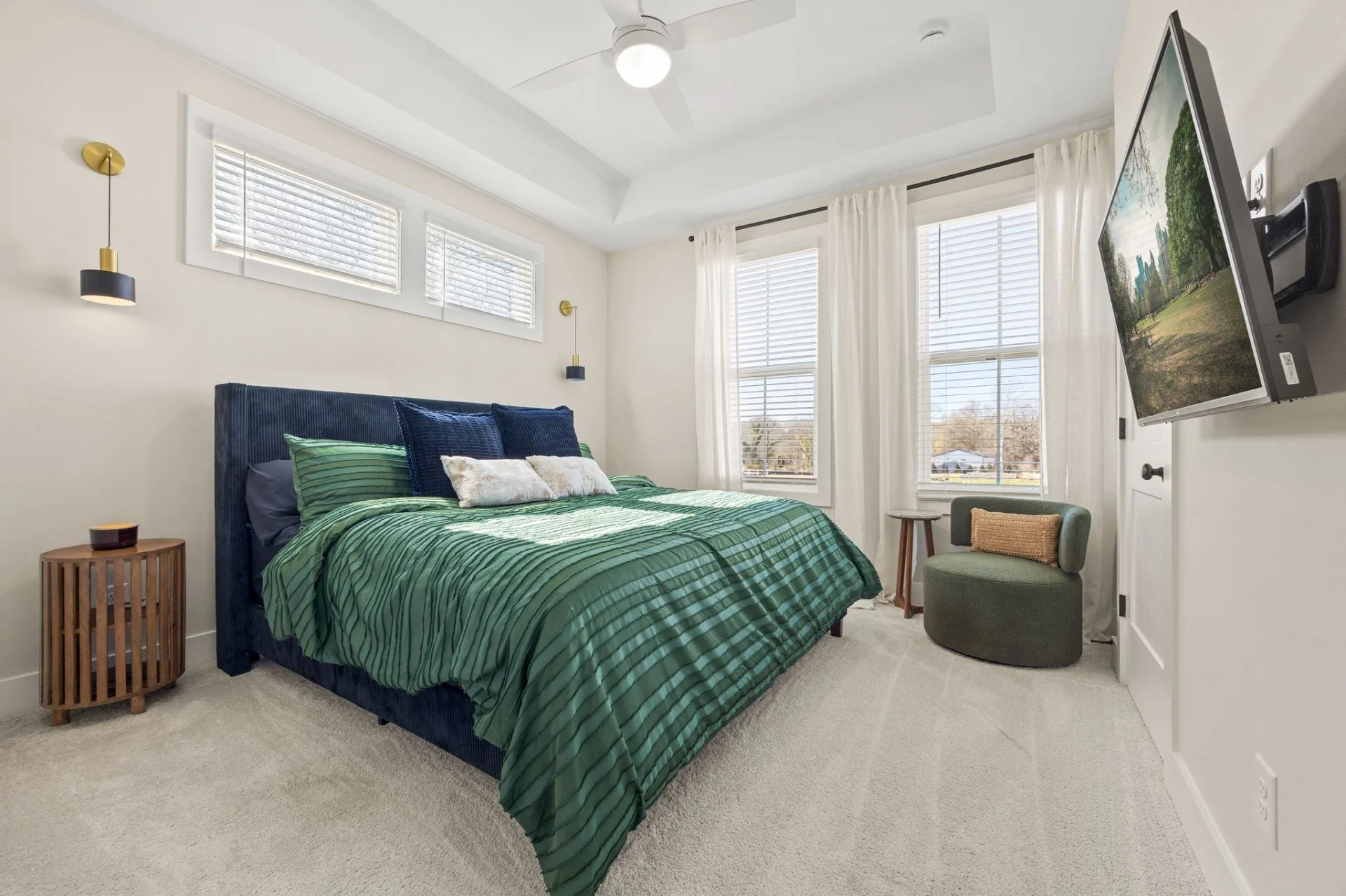 A bedroom with a green bedspread, a blue headboard, and multiple pillows. There are white curtains and windows letting in natural light. A wall-mounted TV is on the right, and there are two wall sconces with black and gold accents on either side of the headboard. A small round side table with a bamboo design is next to the bed, and a green round chair is in the corner.