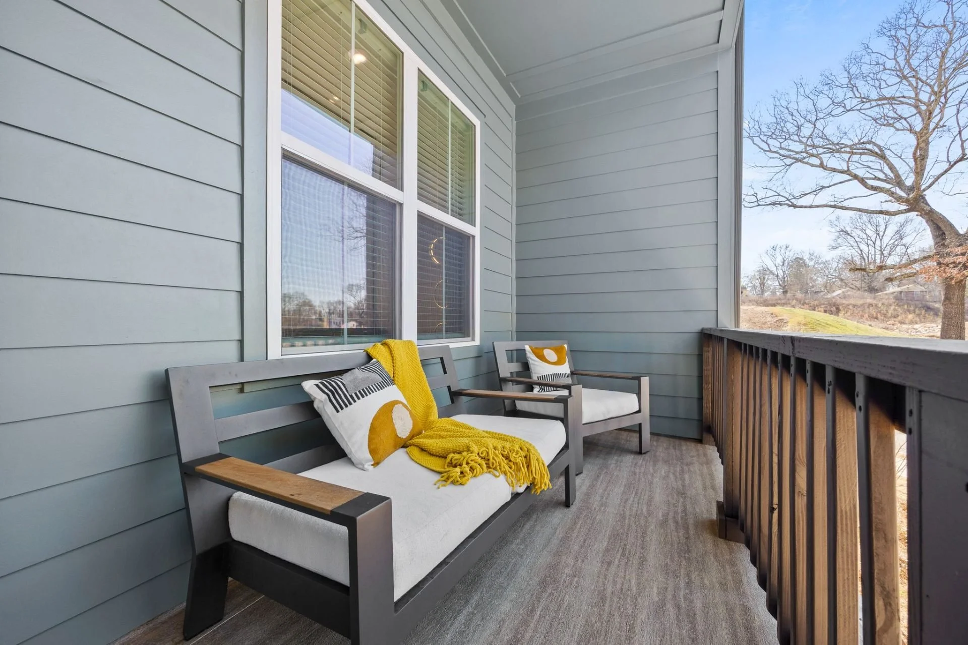 A cozy balcony with two modern chairs featuring white cushions, decorated with black, white, and yellow pillows, and a yellow blanket. The balcony has blue siding, a wooden railing, a gray carpet, and large windows with blinds, overlooking a yard with a large tree and a clear blue sky.