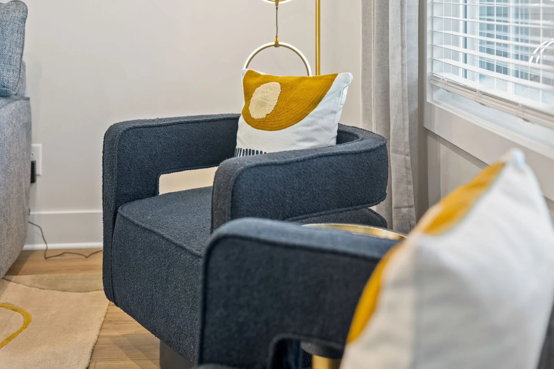 A living room with a navy upholstered armchair, a white pillow with a yellow abstract design, and a window with white blinds. Part of a beige and yellow patterned rug is visible in the corner.