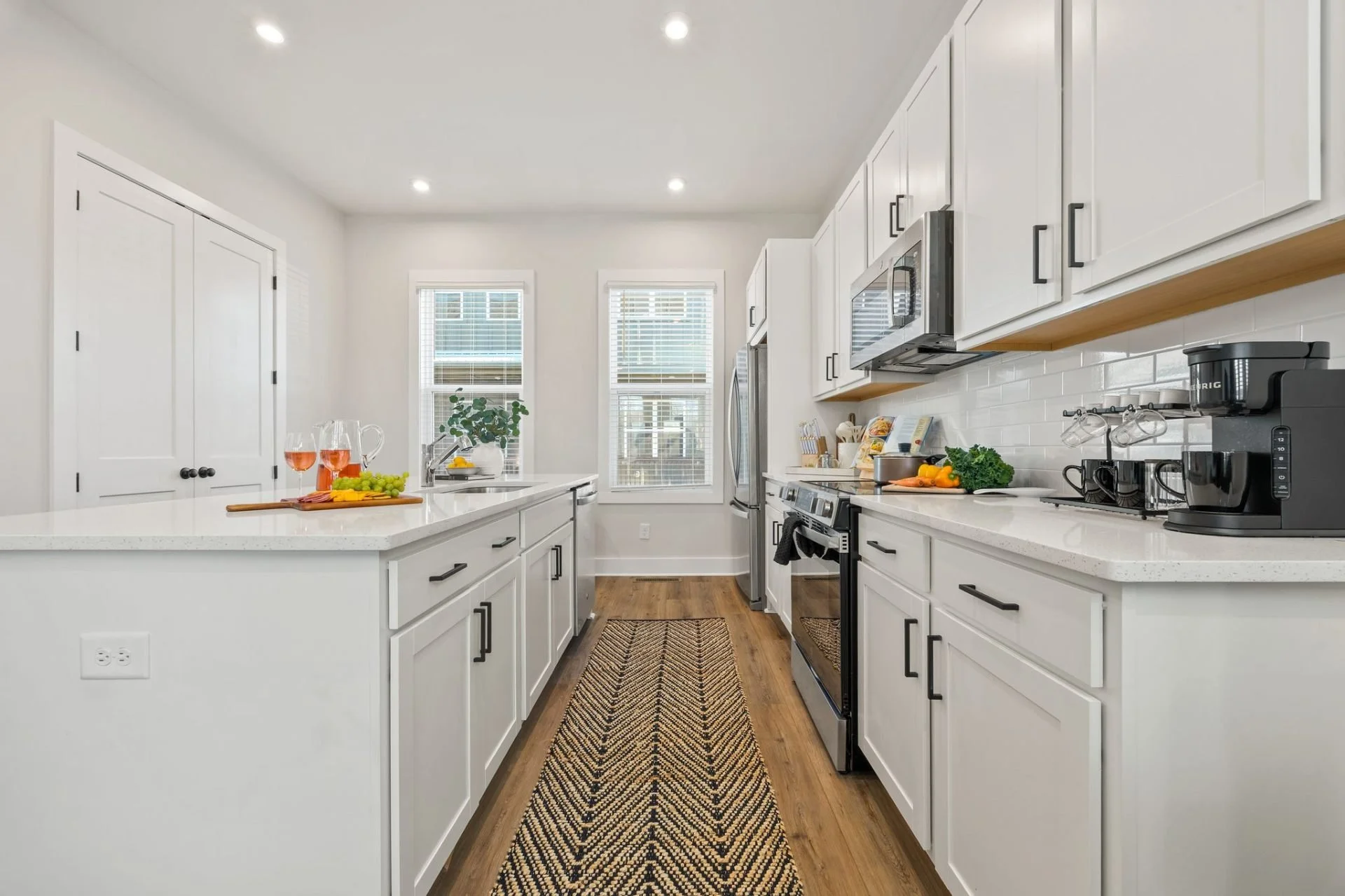 Modern kitchen with white cabinets, stainless steel appliances, a wooden floor, and a long countertop with vegetables, wine glasses, and kitchen gadgets, illuminated by recessed ceiling lights.