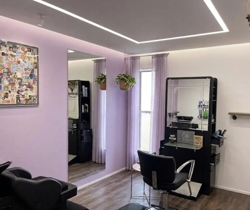 Salon with light purple walls, large mirror and two hanging plants, purple curtains, black styling station with mirror, black styling chair, and a small table. There is a partial view of a black seating area and a colorful picture on the wall.