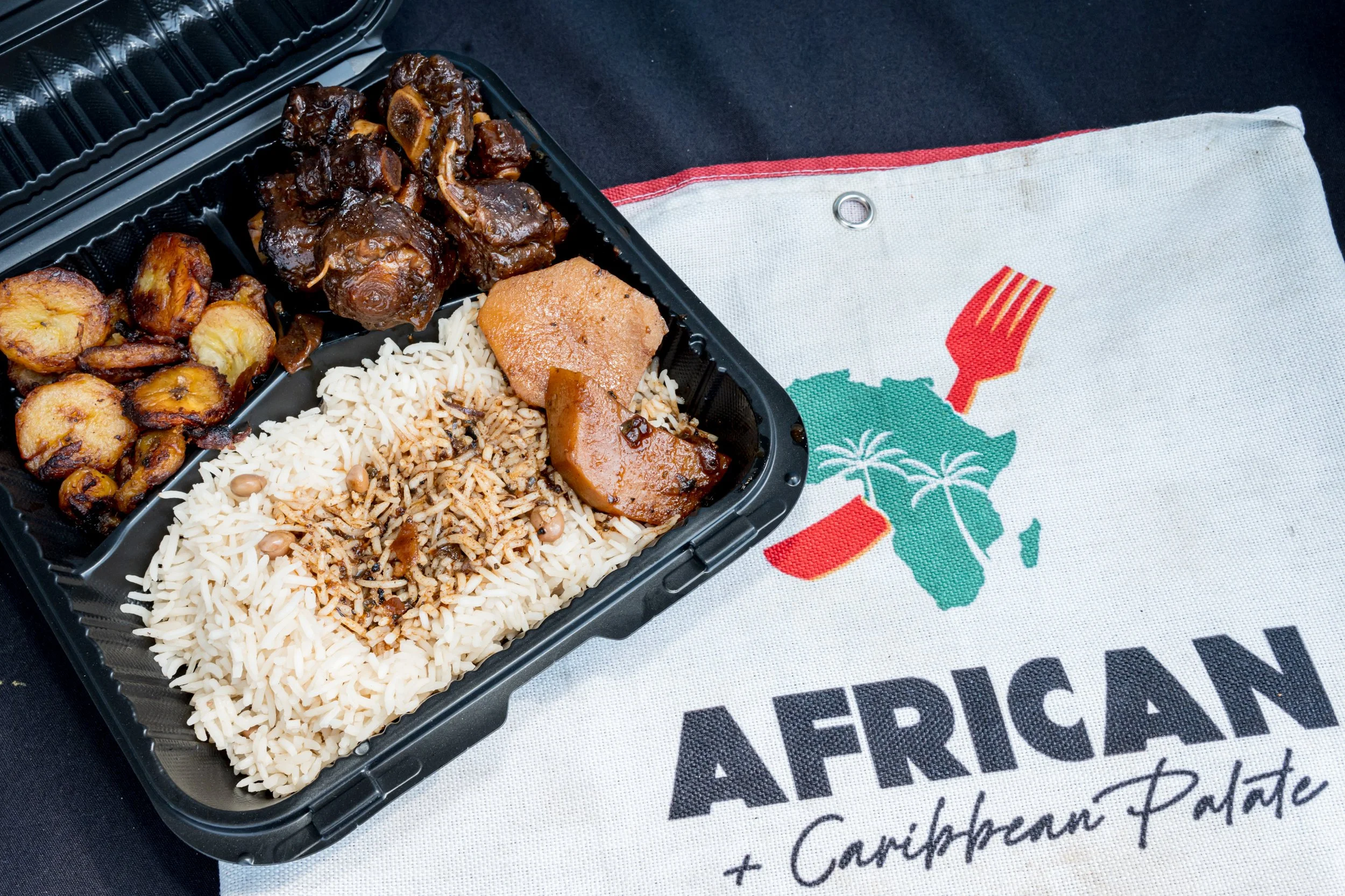 A takeout container with rice, chicken, and plantains, placed on a cloth with a colorful design and text.