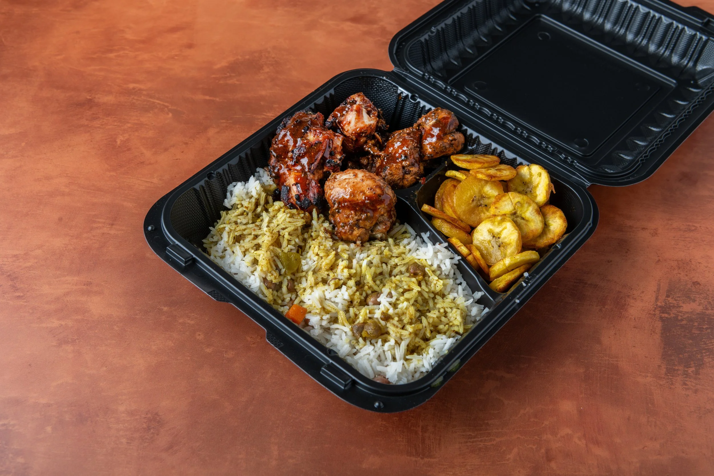A black takeout container with three compartments containing rice, chicken, and plantains on a wooden surface.