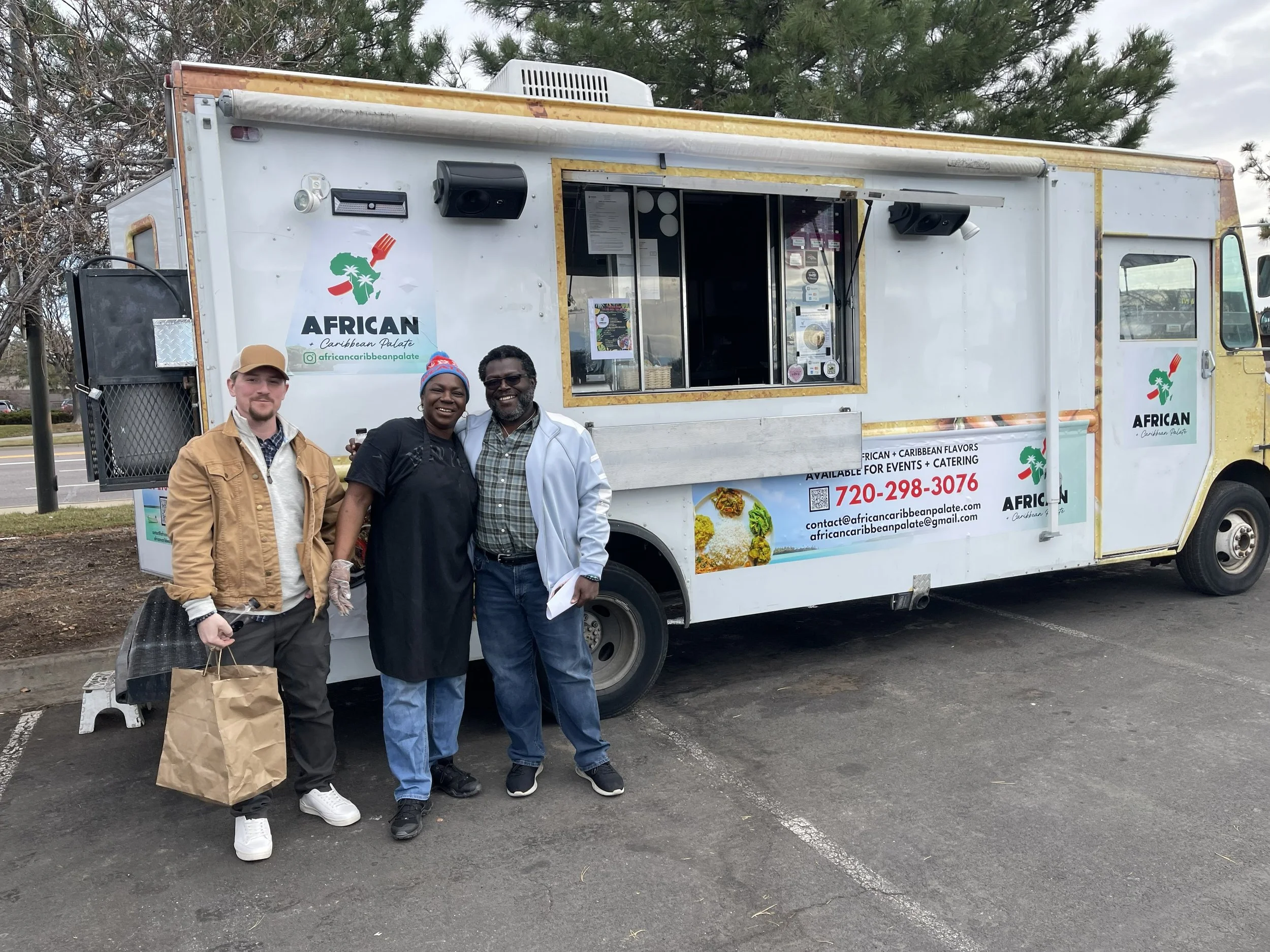 Three people are standing in front of a food truck named African Caribbean Palate. The food truck has a white exterior with logos and contact information displayed. The group includes two men and one woman, smiling and posing for the photo, with the 