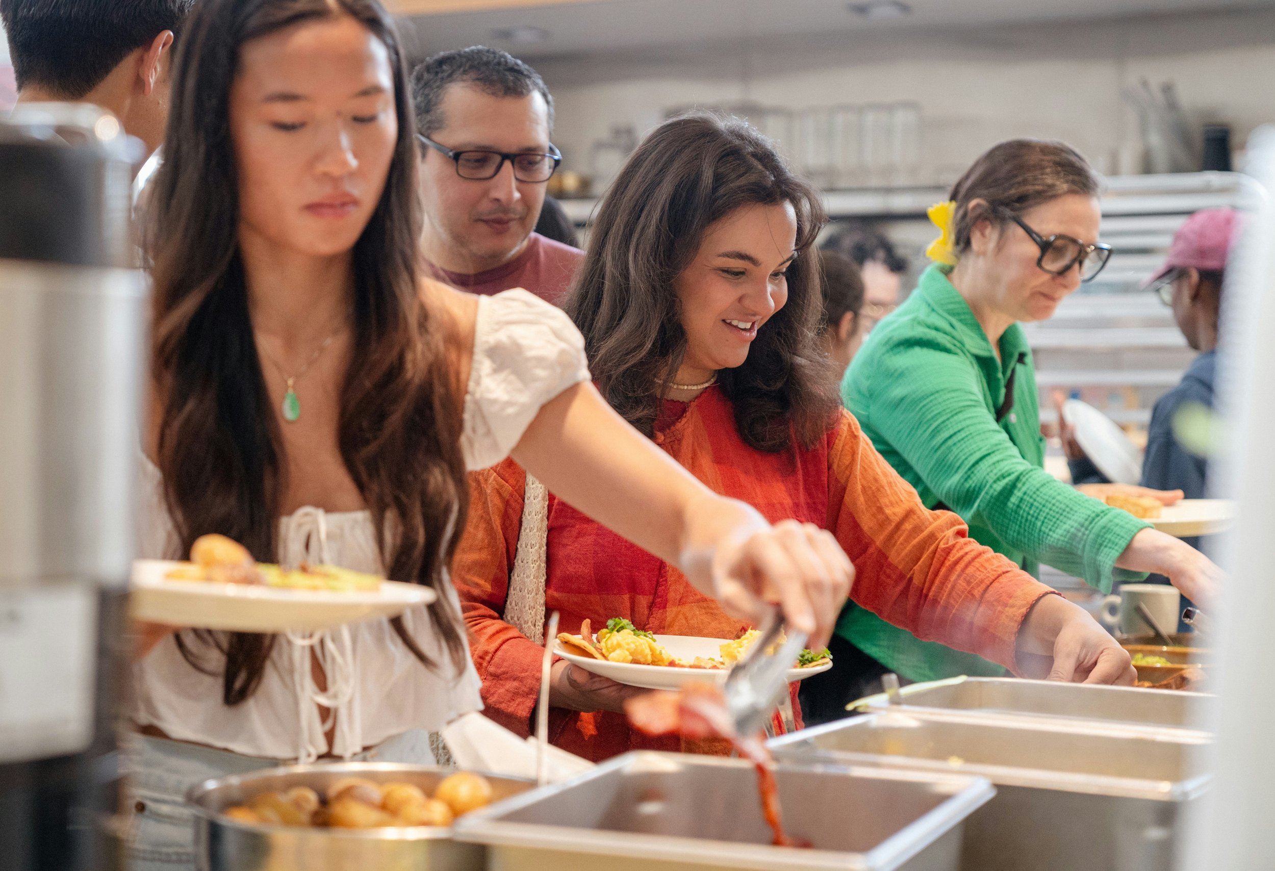 People serving food at a buffet-style restaurant or event, with various dishes and food trays in the foreground and background.