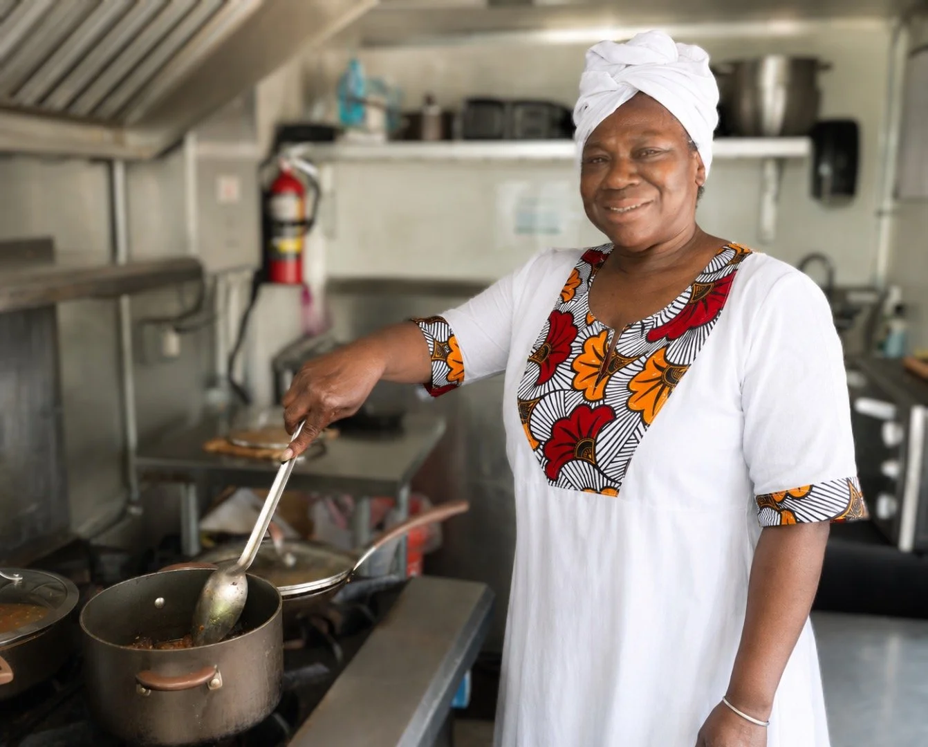 A woman cooking on a stove in a kitchen, wearing a white dress with colorful floral patterns and a white headwrap, smiling while stirring a pot.