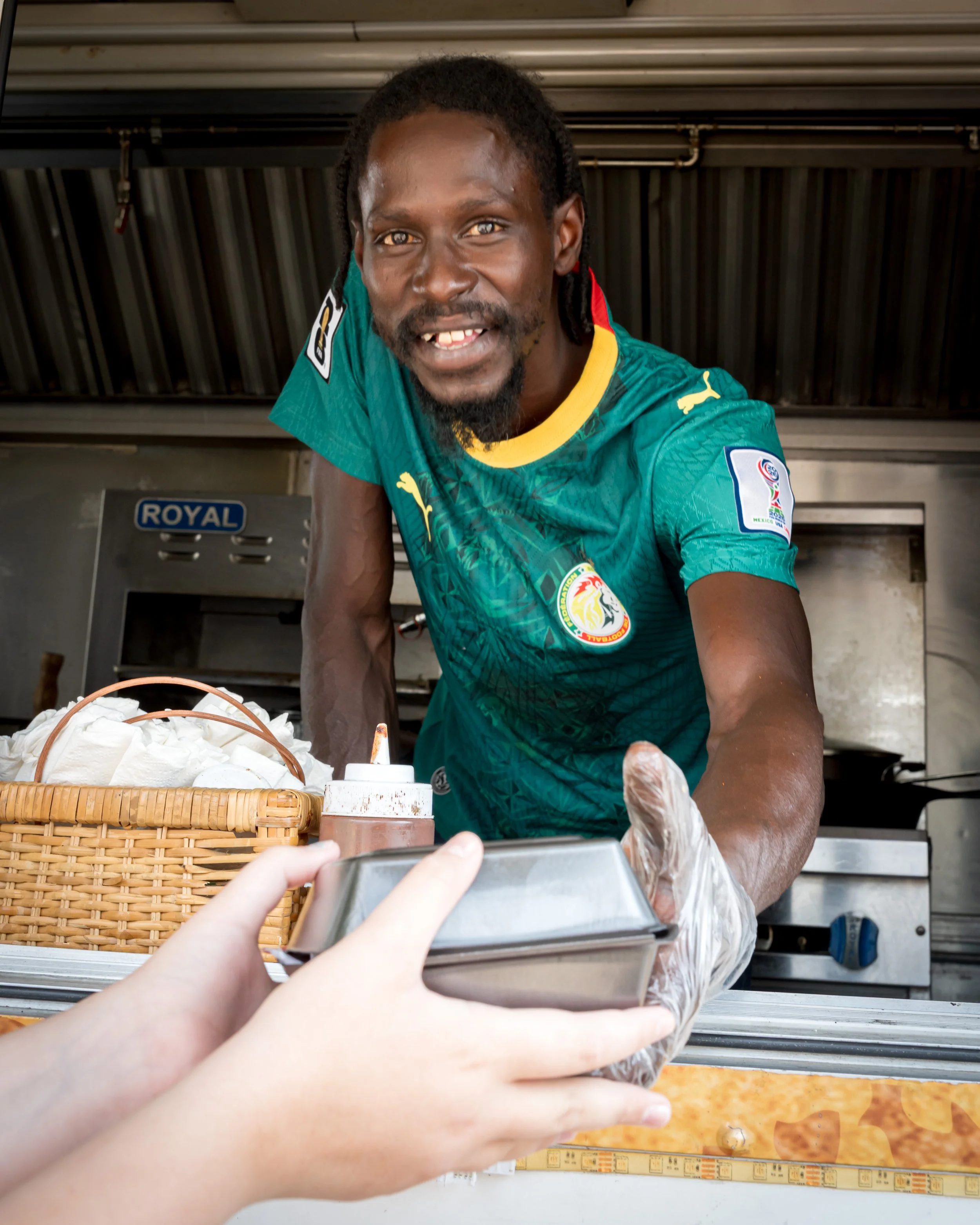 A man, wearing a green soccer jersey with a yellow collar, is smiling at the camera while leaning  counter. He appears to be working at a food stand or kitchen, with a basket of bread and condiment bottles in front of him. There is a person in the fo