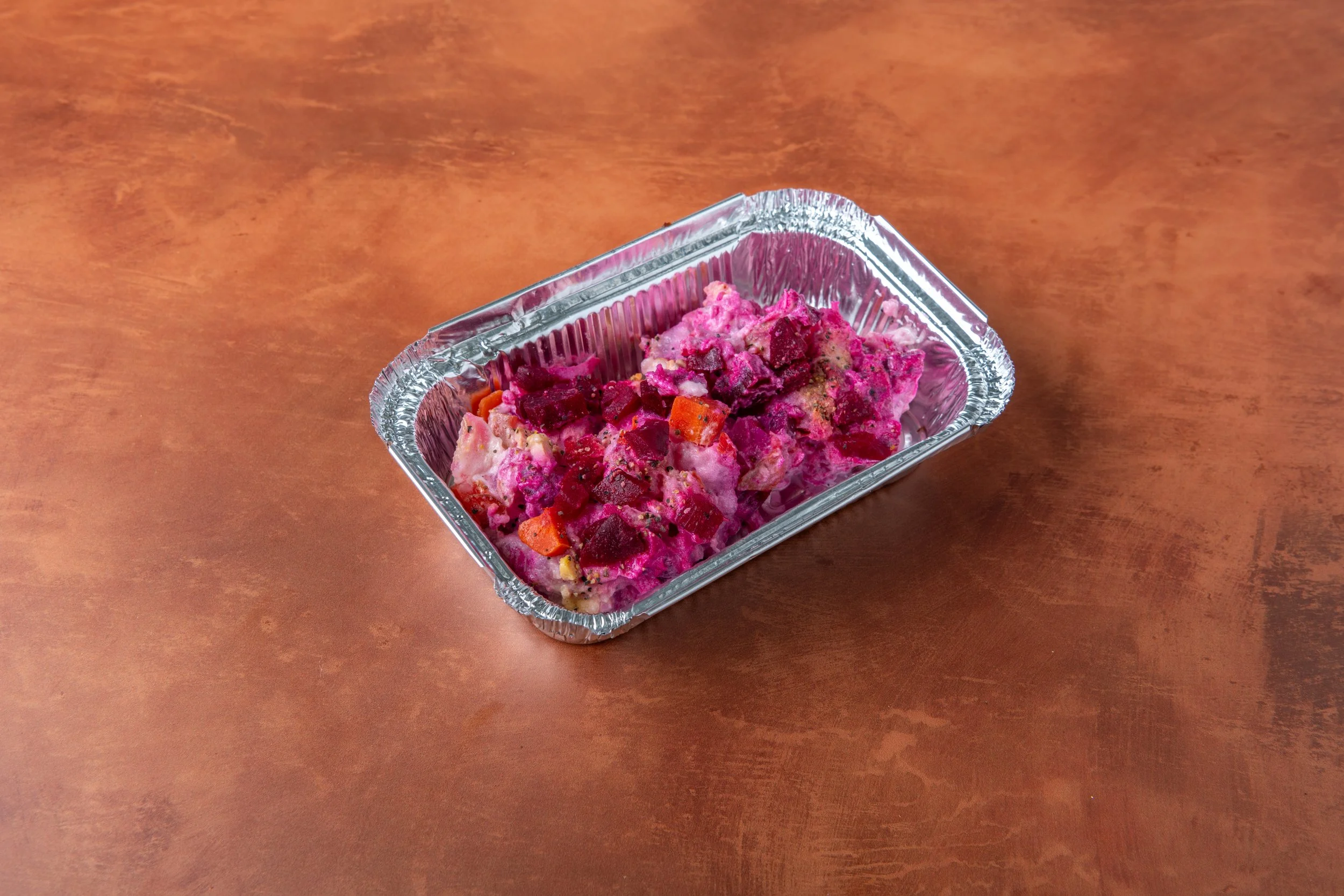 A small aluminum tray of creamy potato salad with diced potatoes, vibrant beets, and carrots, blended with a tangy dressing on a wooden surface.