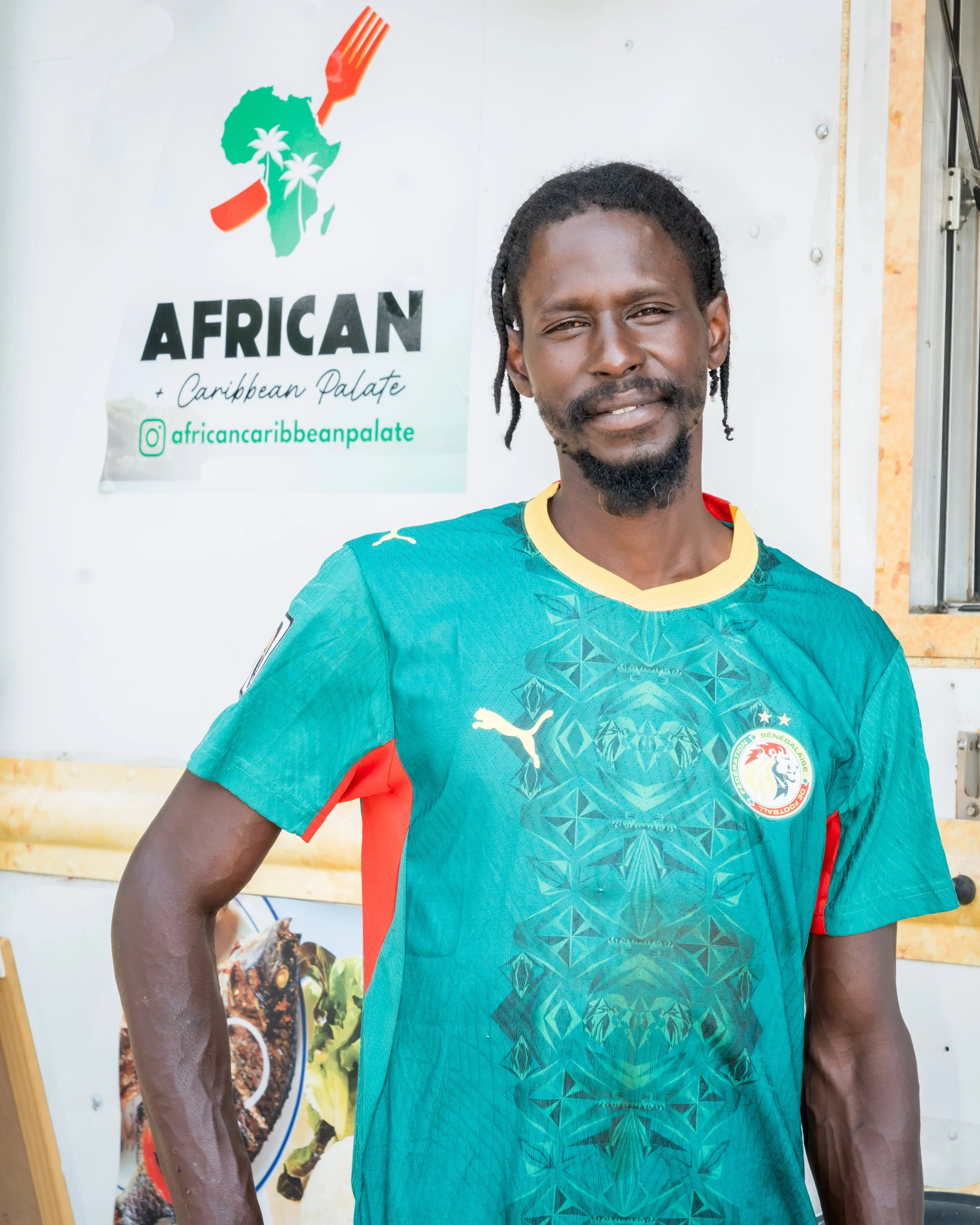 A man with dark skin, dreadlocks, and a beard, wearing a green and red sports jersey, standing in front of a white background with a poster that reads 'African + Caribbean Palate' and features a map of Africa with palm trees, and a social media handle @africancaribbeanpalate.