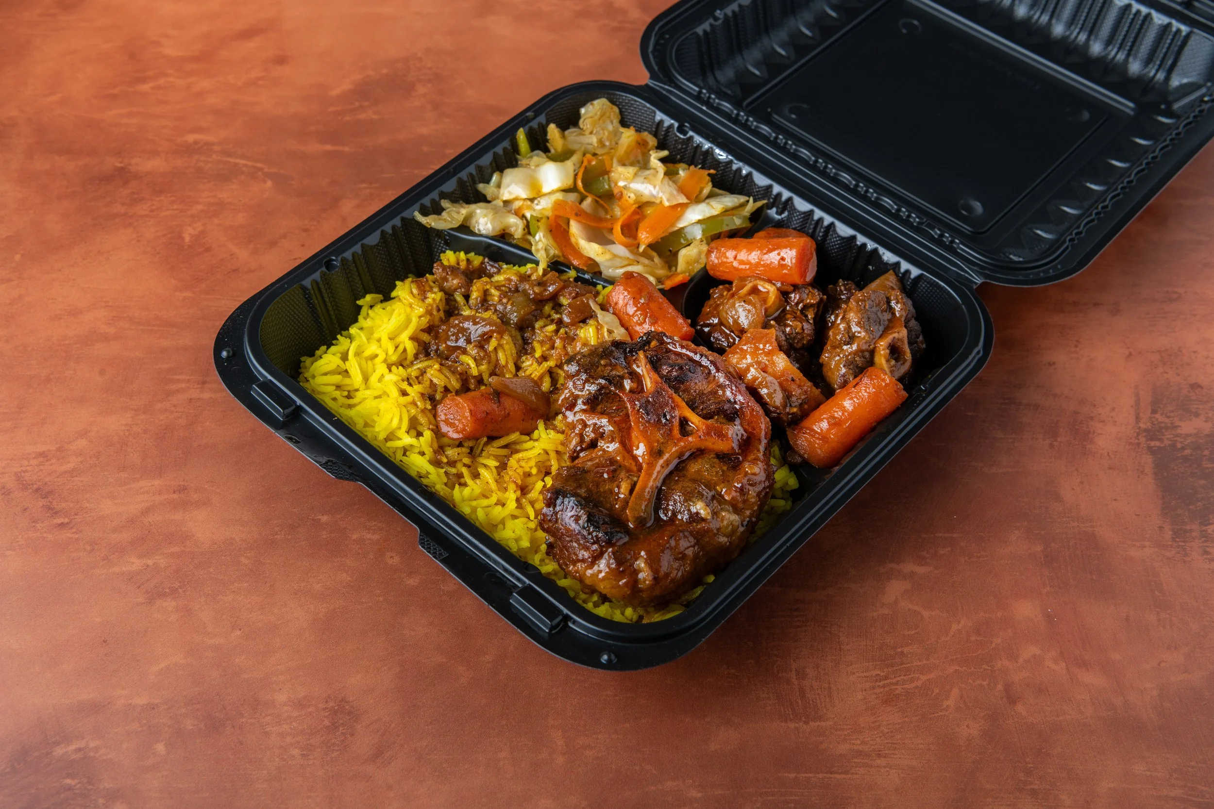 A black plastic takeout container with compartments holding yellow rice, beef stew, cooked vegetables, and a mix of stir-fried vegetables including cabbage, carrots, and peppers, on a brown surface.