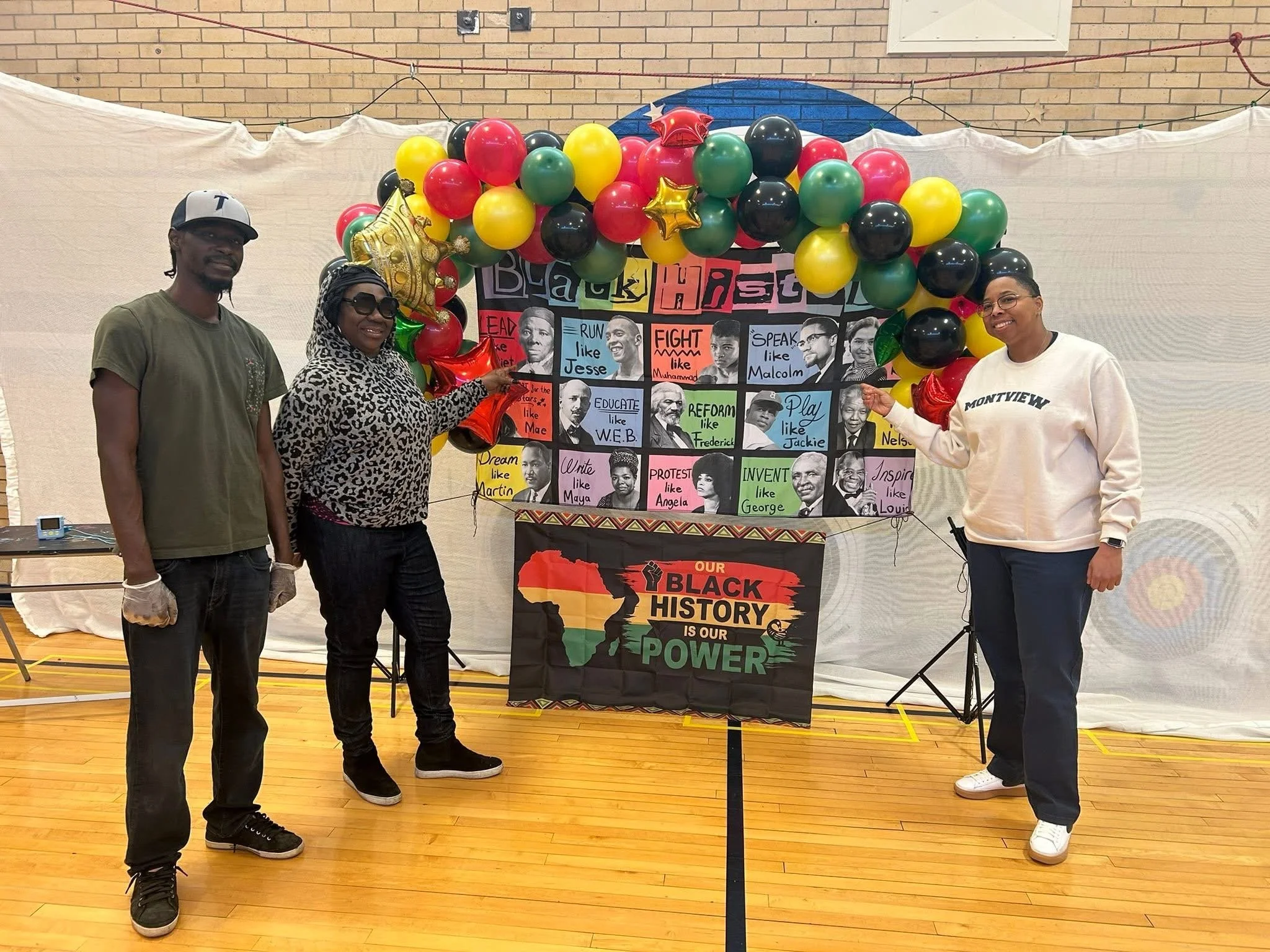 Three people standing in front of a colorful display with balloons and photographs, holding pointed tools, in a gymnasium with wooden floors and exposed brick wall.