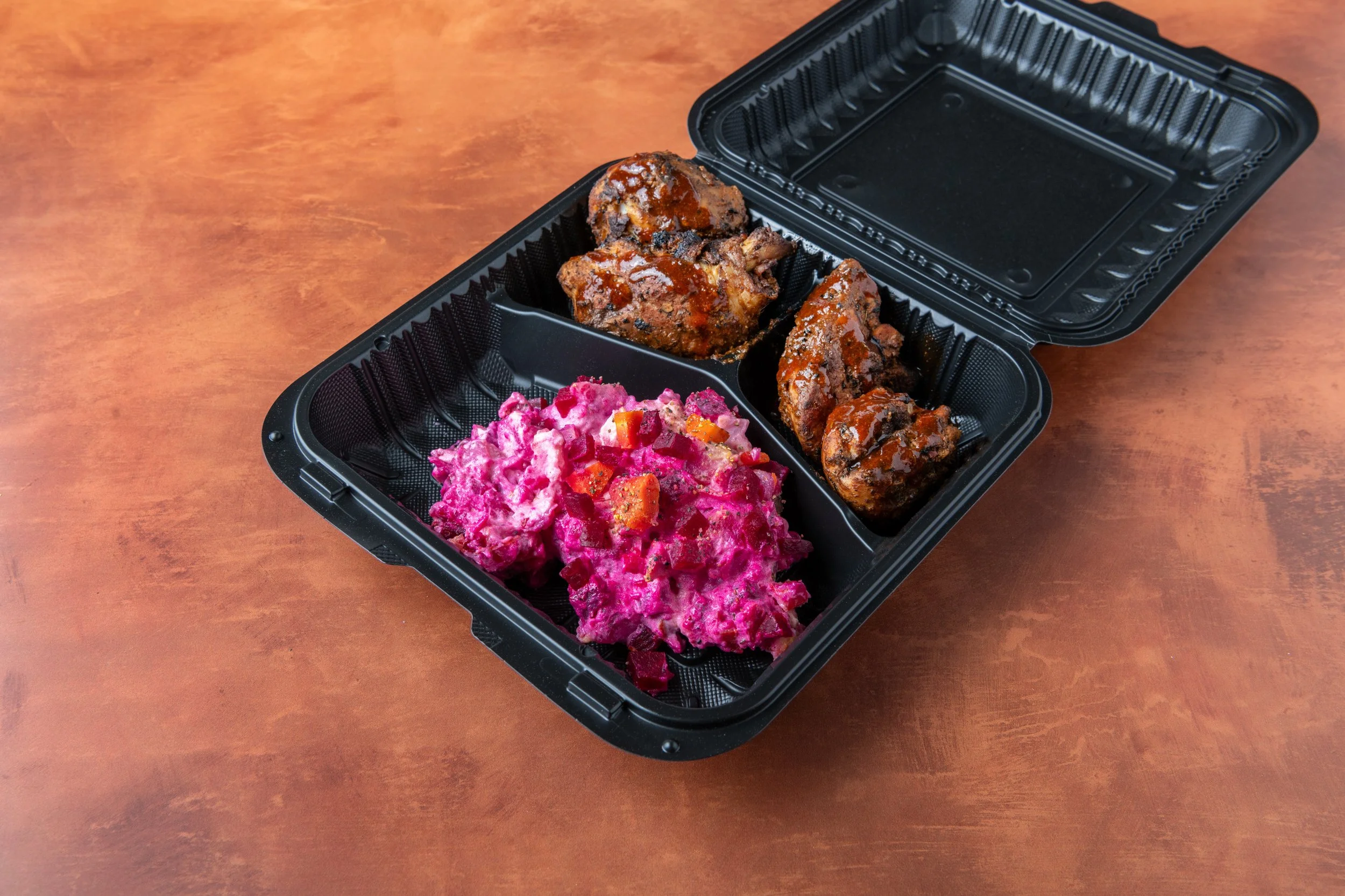 A black takeout container holding three pieces of glazed meat and a serving of beet salad on a wooden surface.