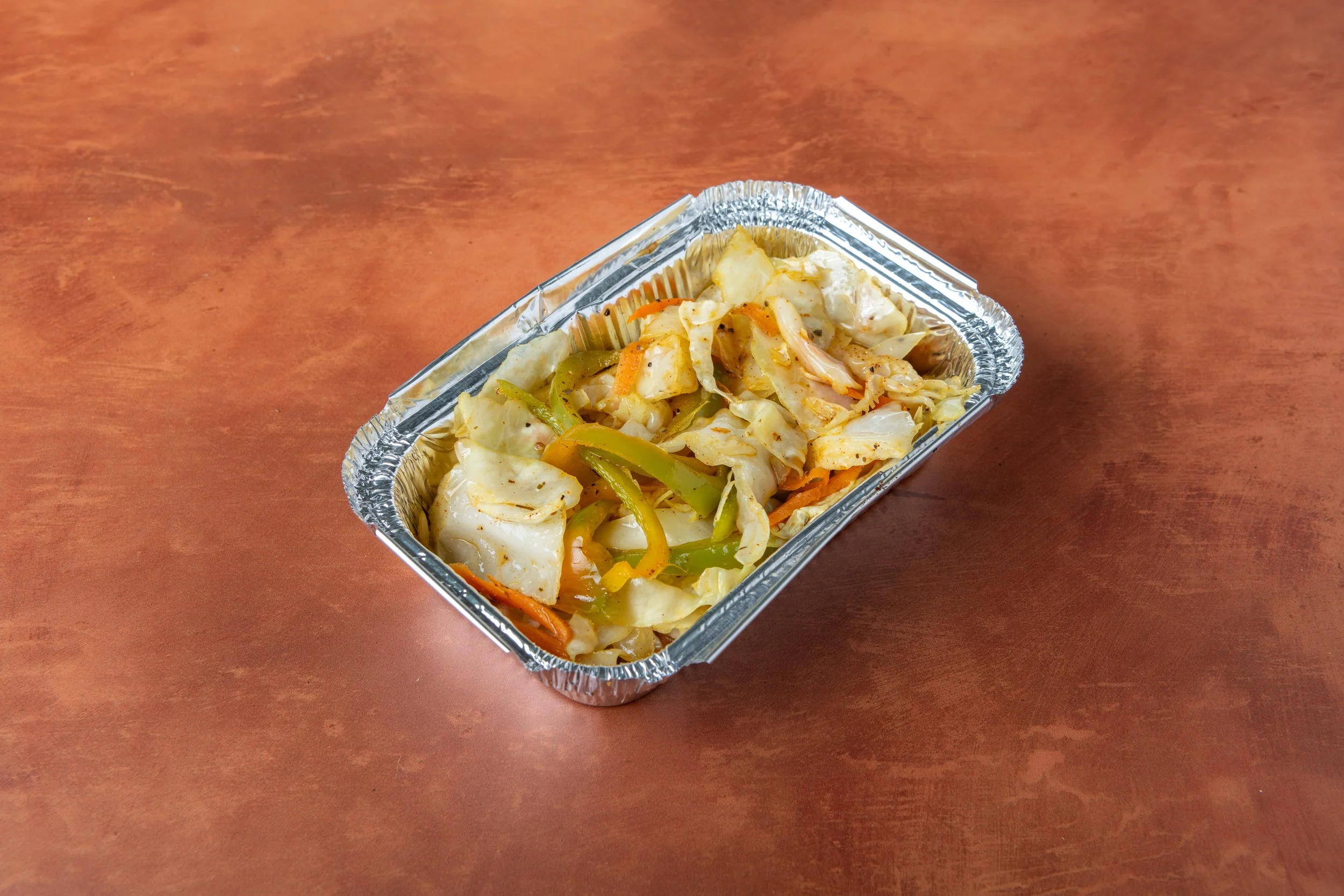 A rectangular aluminum tray containing cooked cabbage, bell peppers, and carrots, placed on a reddish-brown wooden surface.