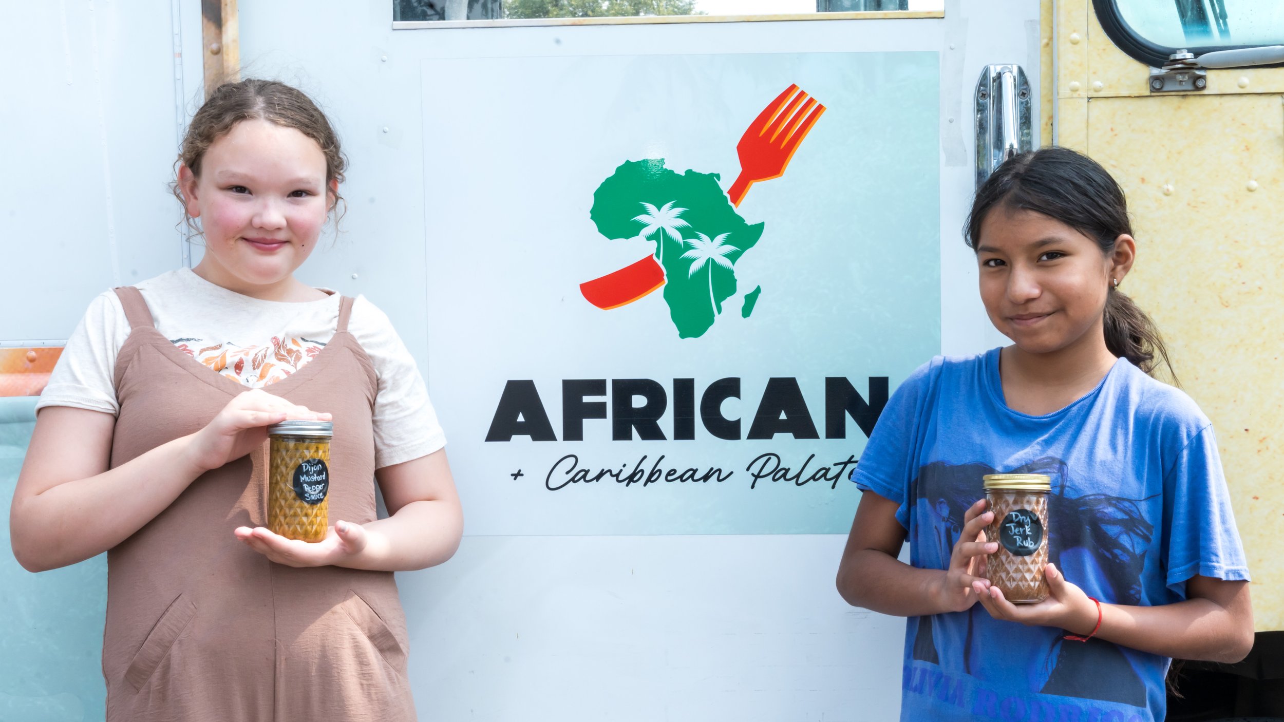 Two young girls holding jars of sauce, standing in front of food truck, with the text "African + Caribbean Palate."