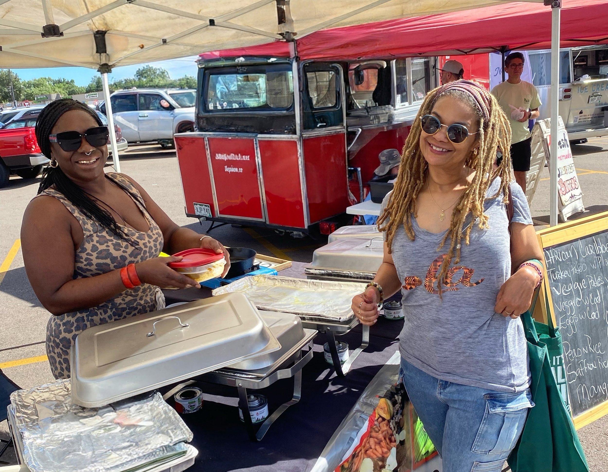 Two smiling women, one with dark skin and sunglasses, the other with light skin and dreadlocks, standing at a food stall in a parking lot on a sunny day.