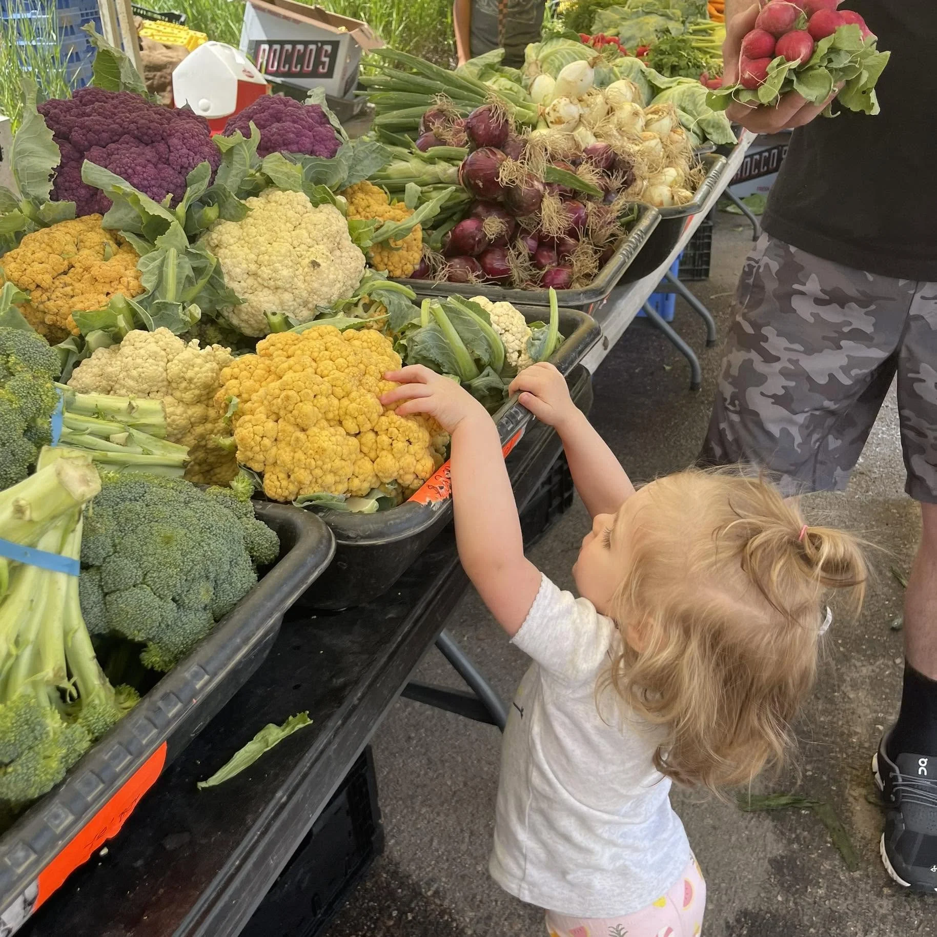 A young girl with curly blonde hair reaching for yellow and purple cauliflower at a farmer's market stand with various vegetables like onions, radishes, and broccoli displayed.