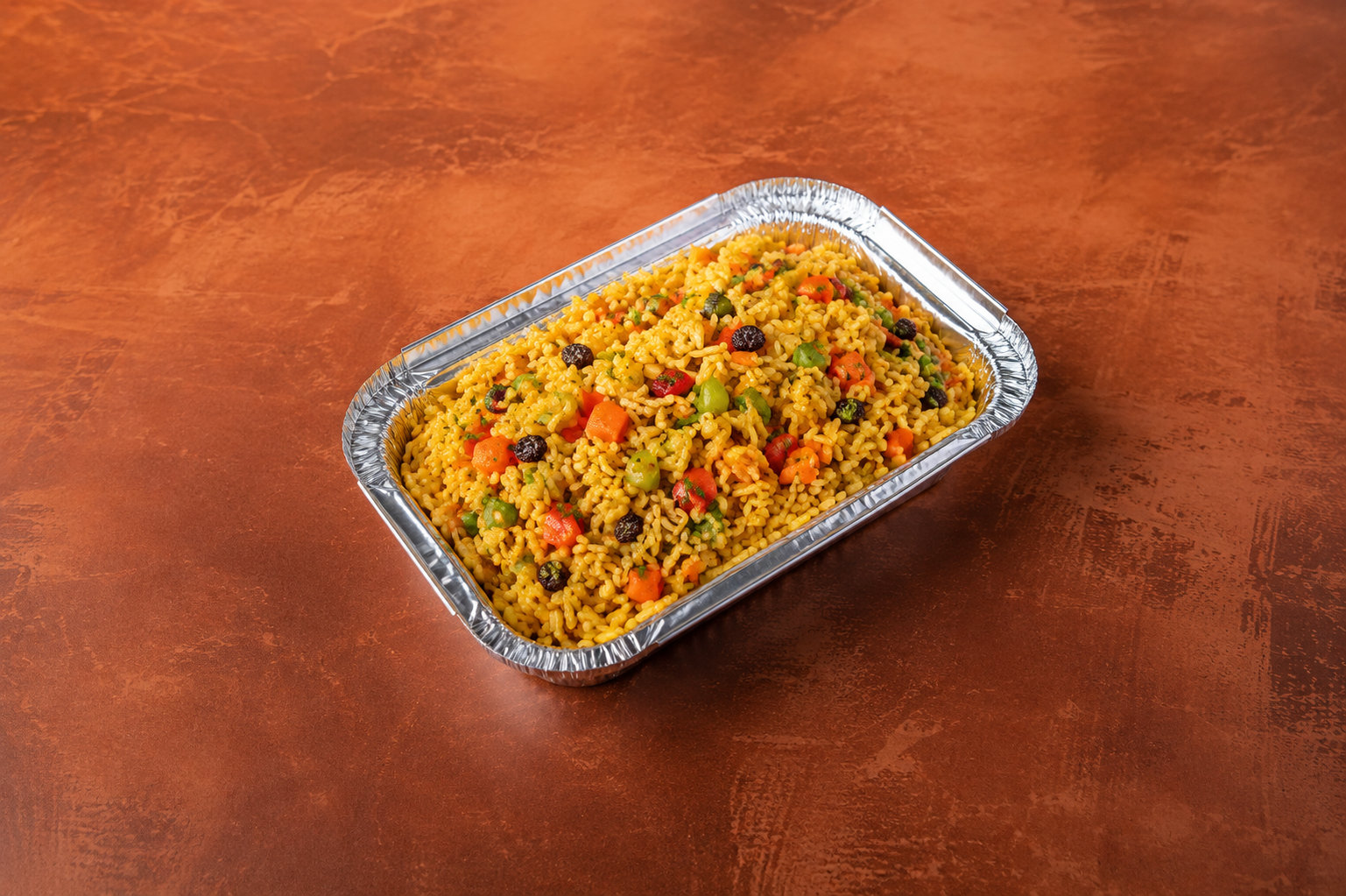 A foil tray filled with seasoned yellow rice mixed with diced vegetables and raisins, placed on a reddish-brown textured surface.