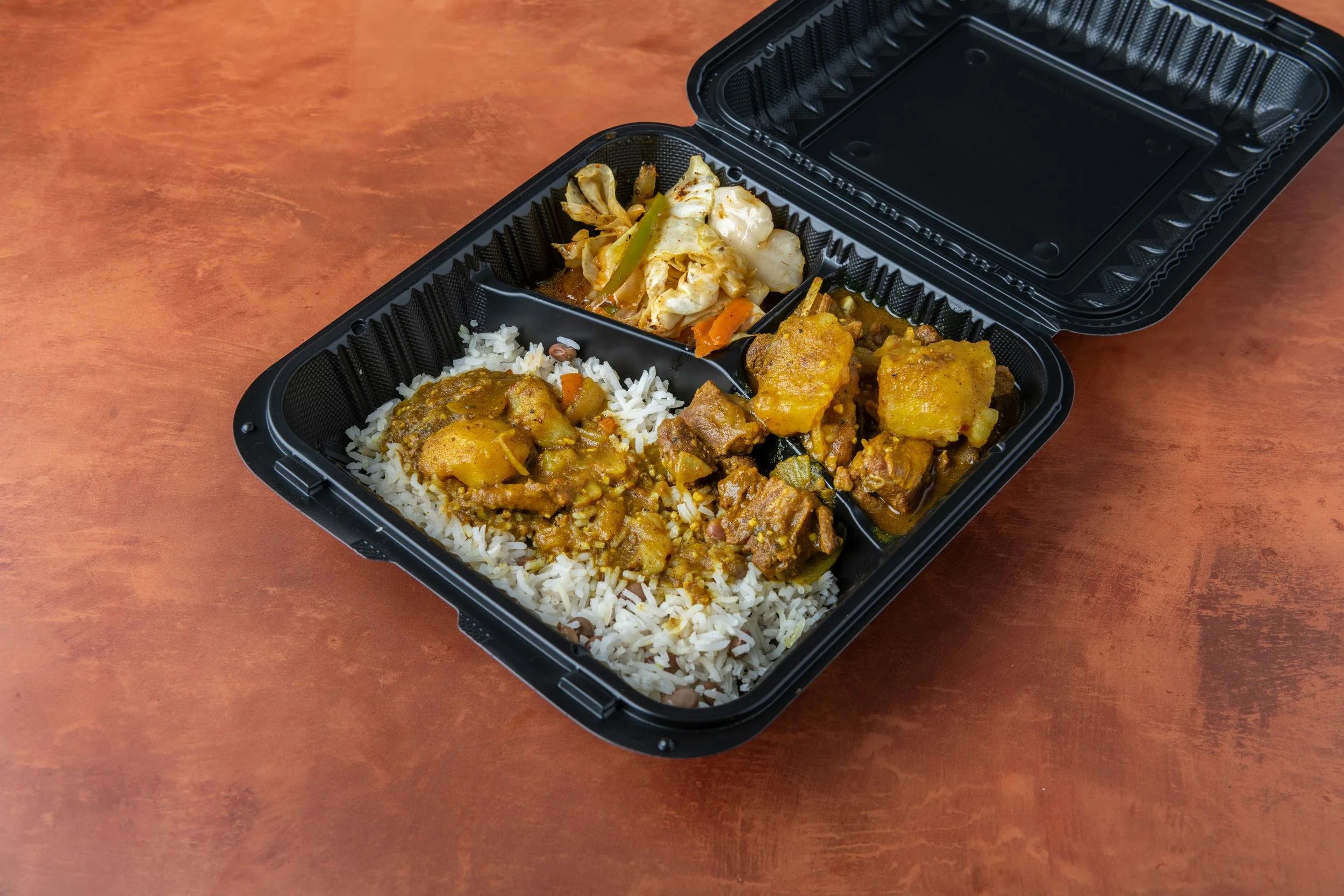 A black plastic takeout container with rice and curry and vegetables inside. The container is on a wooden table.