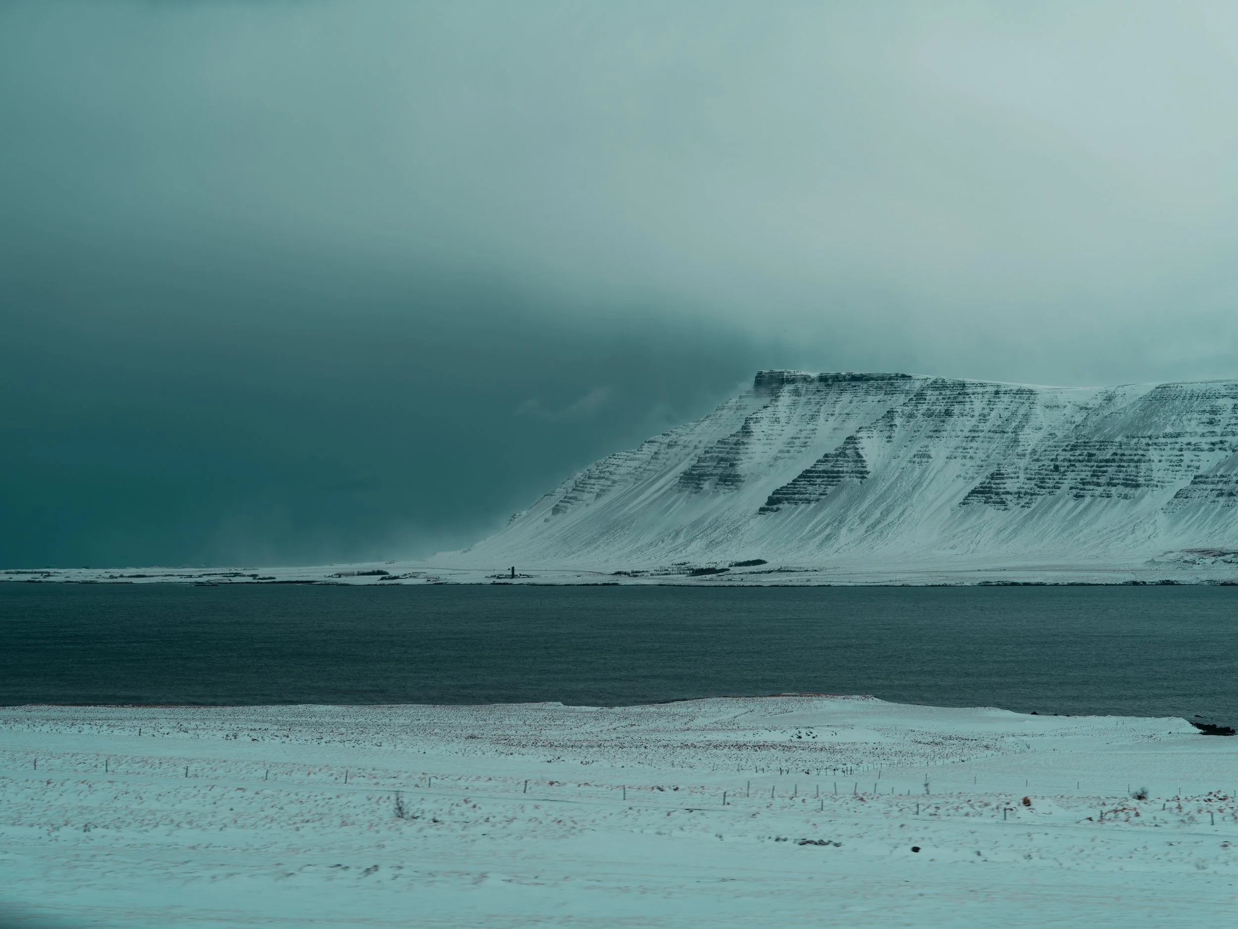 Snow-covered landscape with a mountain in the background and a large body of water in the foreground.