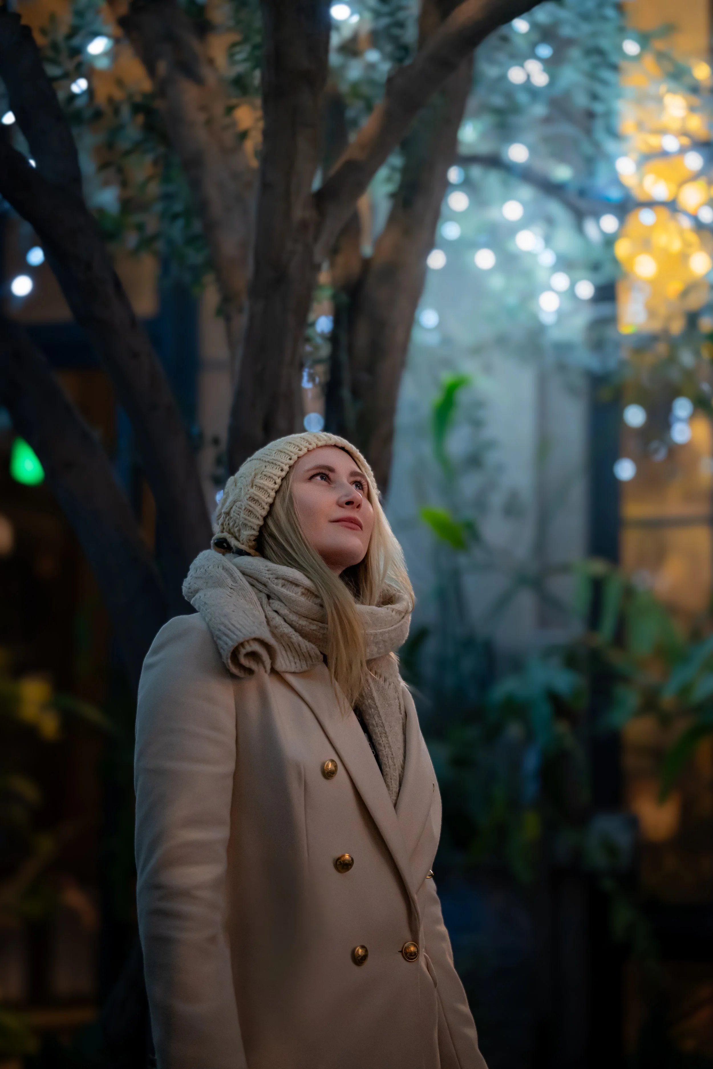 A woman wearing a beige coat and knit hat looking up outdoors with trees and blurred lights in the background