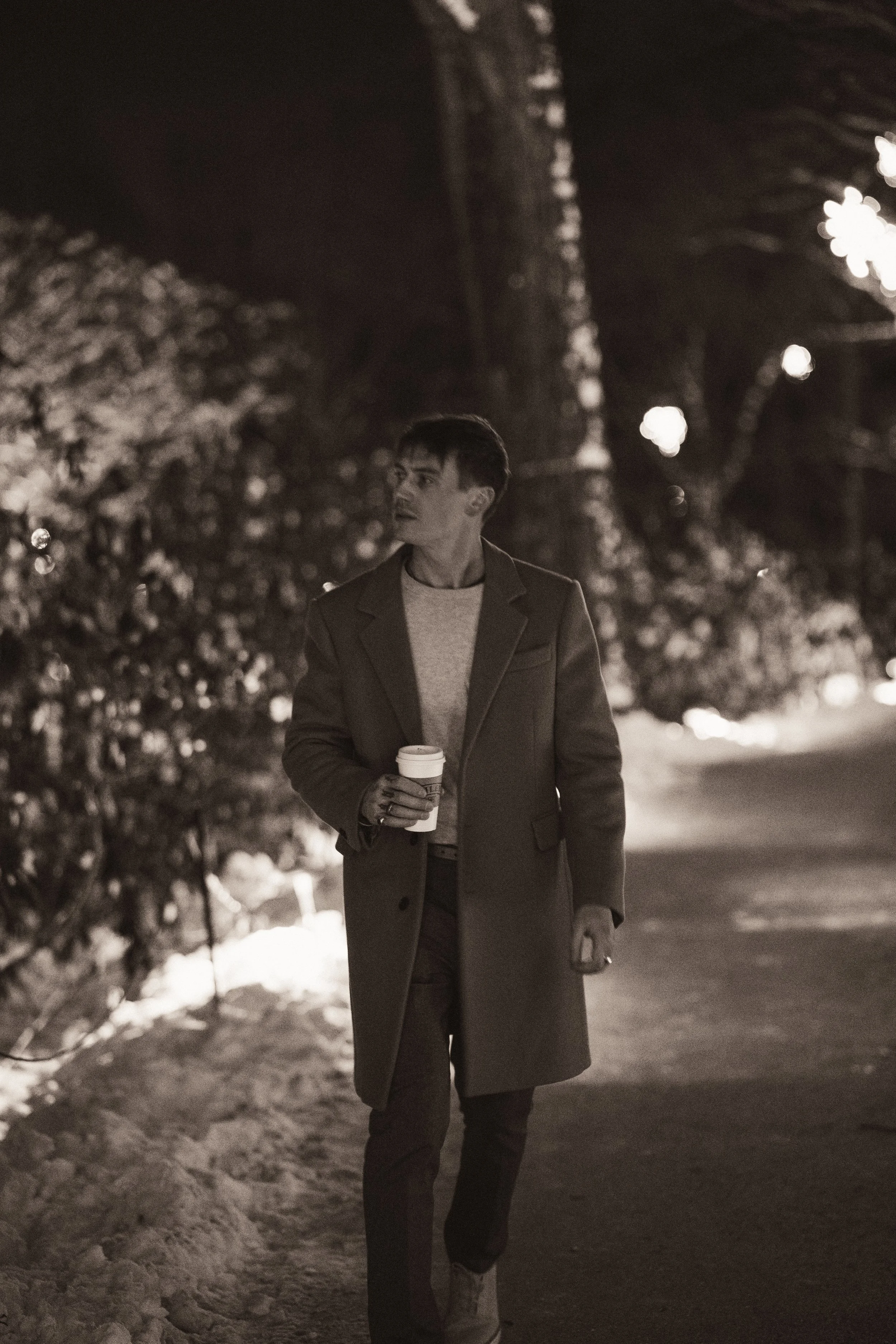 A young man walking outside at night, holding a coffee cup, wearing a coat, in a snowy area with trees and streetlights in the background.