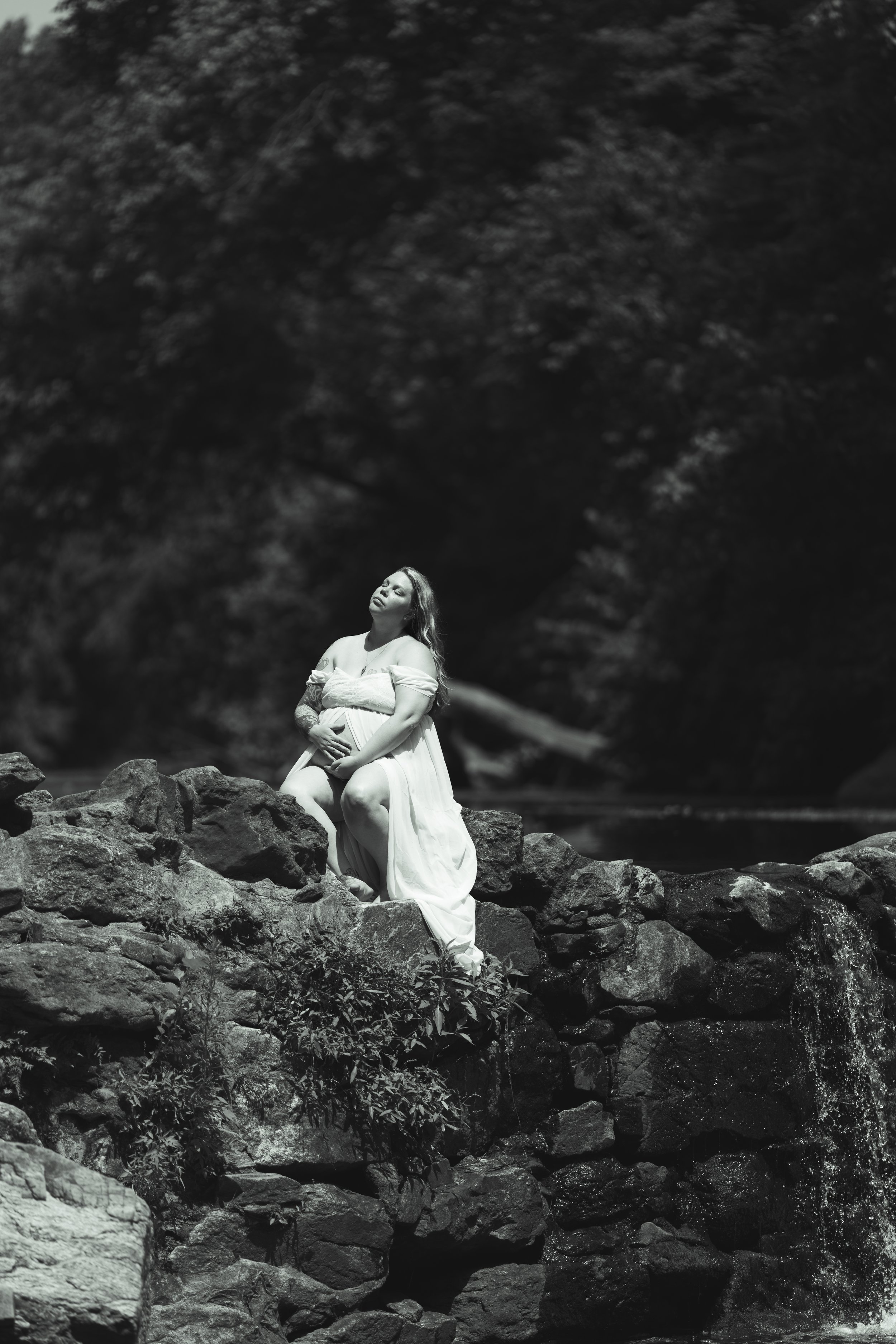 A woman in a flowing dress sitting on rocks near a body of water in a natural outdoor setting, with trees and foliage in the background, in black and white.