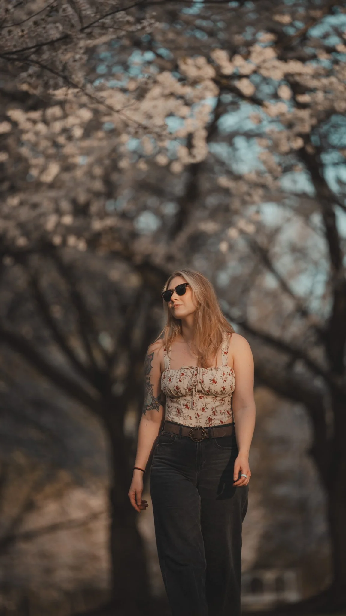A woman with long hair and sunglasses standing outdoors among blooming trees with white flowers, wearing a floral sleeveless top and dark jeans.