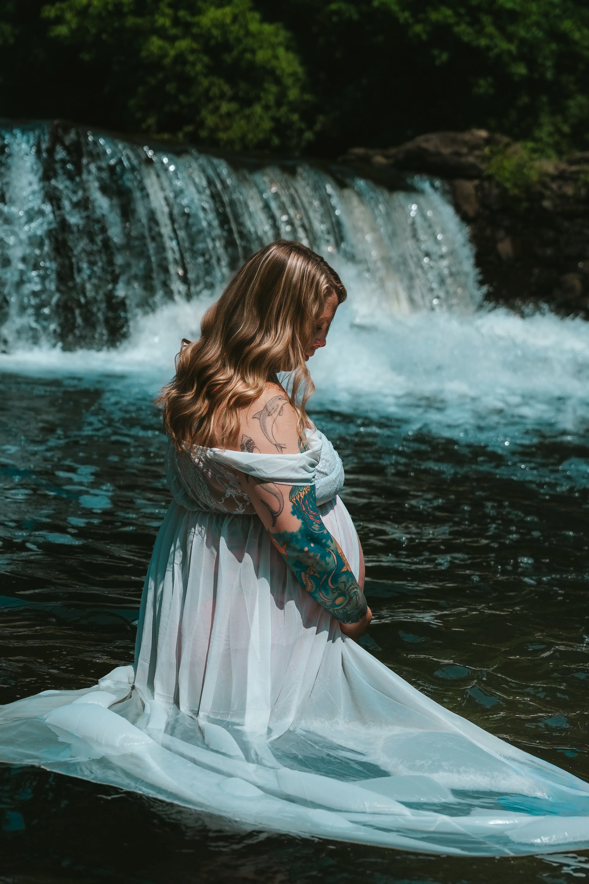 A woman with long wavy hair, tattoos on her arms, wearing a flowing white dress, standing in a river near a waterfall, with lush green trees in the background.