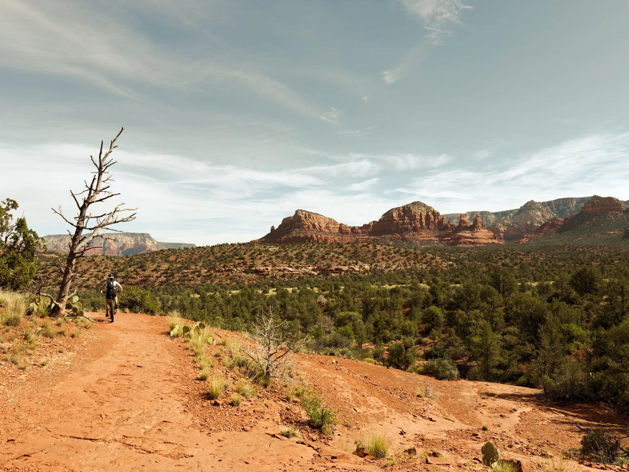 A hiker on a dirt trail in a desert landscape with red rock formations and sparse vegetation under a partly cloudy sky.