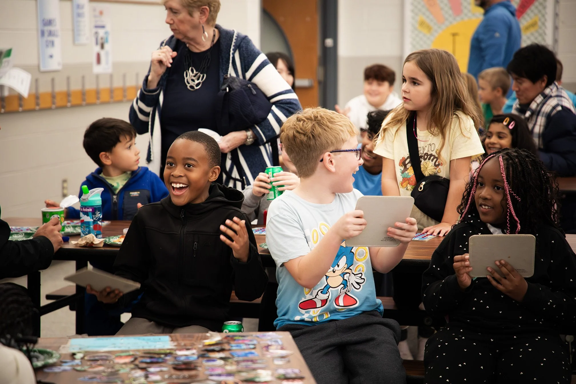 A diverse group of children and a teacher in a classroom, engaging in activities. The children are smiling and holding tablets, with snacks and drinks on the tables.