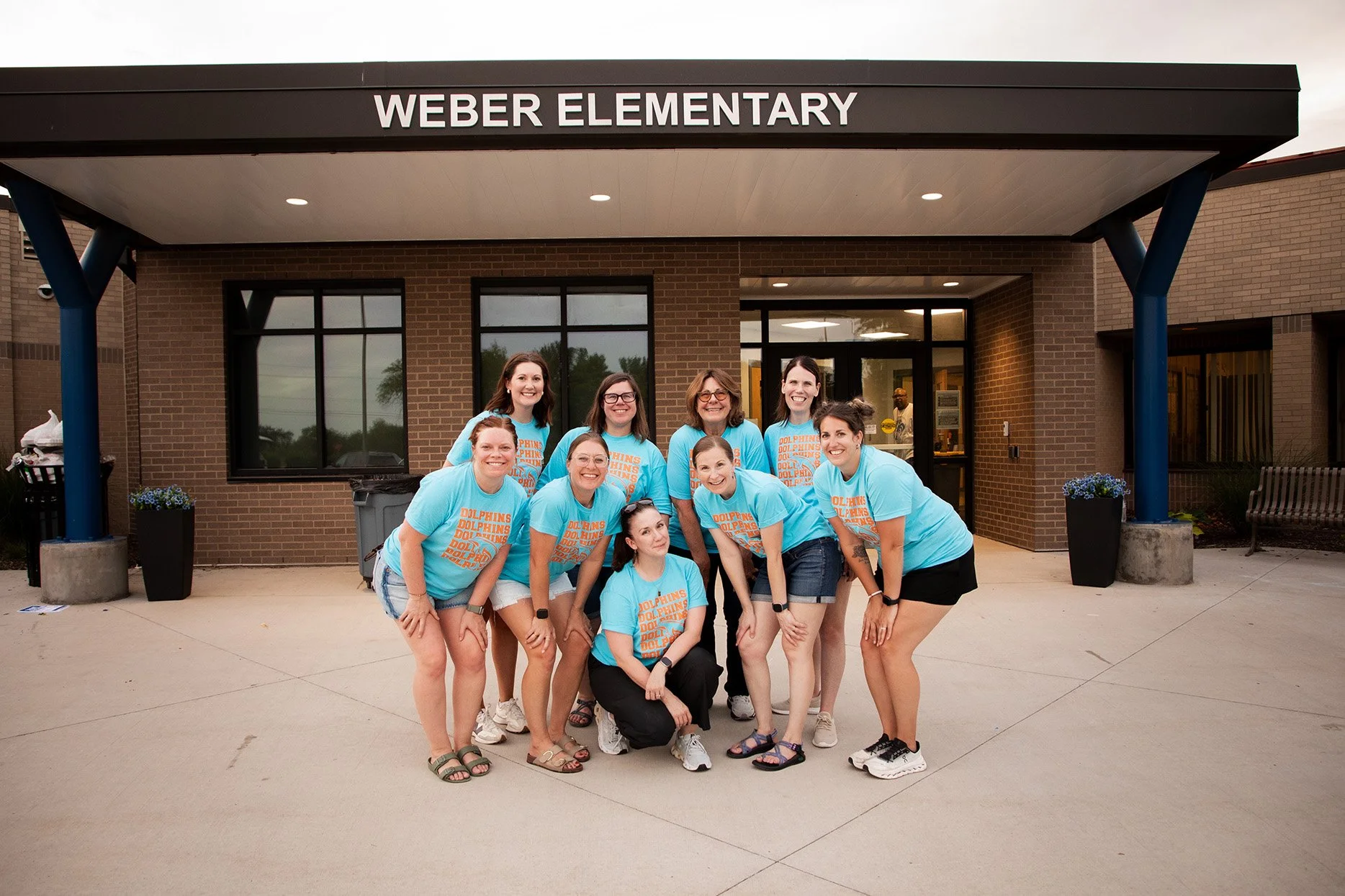 A group of women in matching turquoise shirts with orange text, posing in front of Weber Elementary school building.