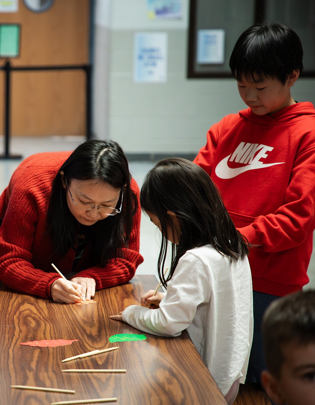 An adult woman and two children engaging in a craft activity at a wooden table, with colorful paper and sticks on the table in a classroom or community center setting.