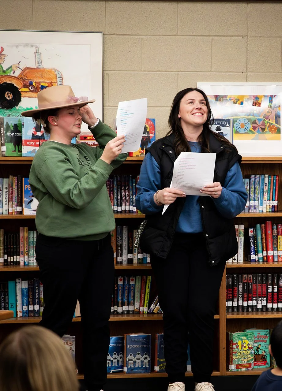 Two women standing in front of a bookshelf in a library, holding papers. The woman on the left wears a wide-brimmed hat and a green sweatshirt, adjusting her hat. The woman on the right is smiling, wearing a blue shirt with a black vest. Others are seated in front of them, partially visible.