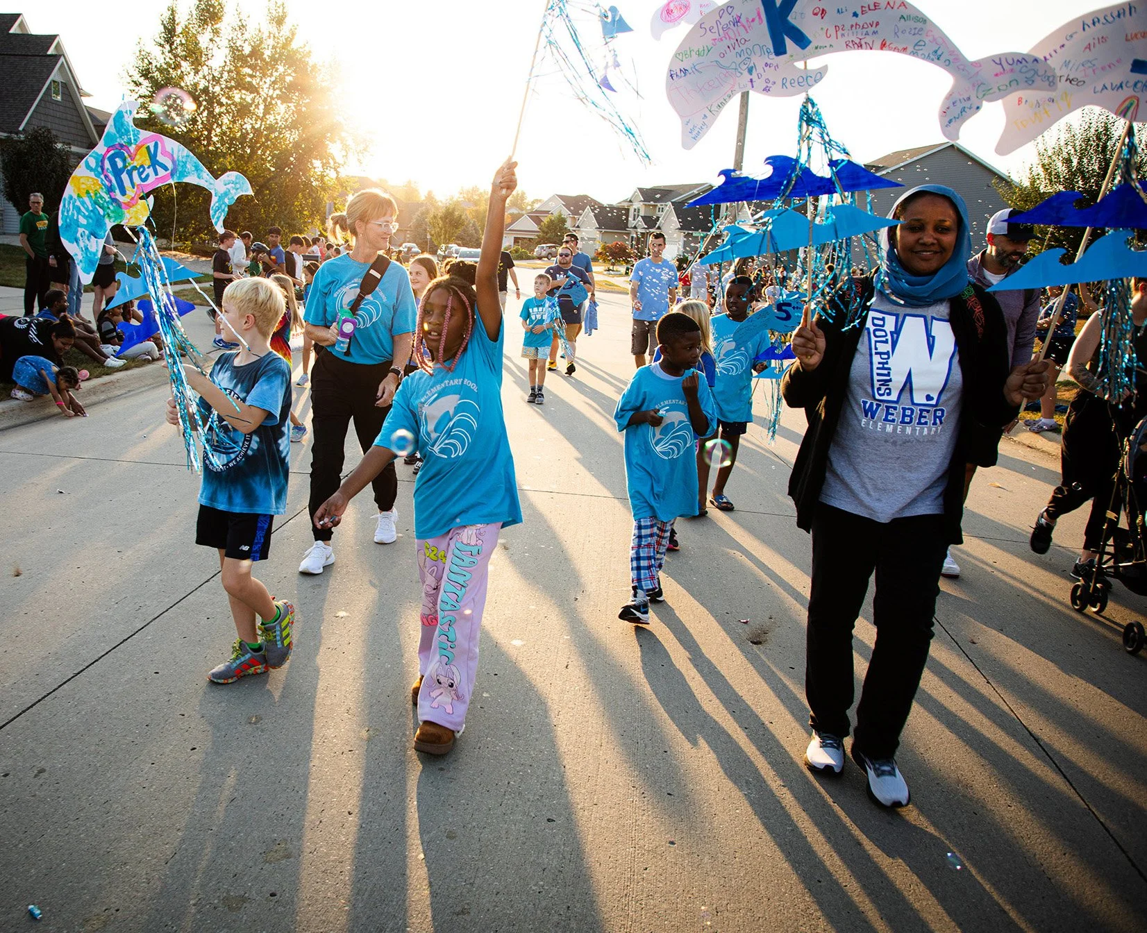 Children and adults participating in a parade on a residential street, holding colorful decorations and signs, with the sun setting behind trees.