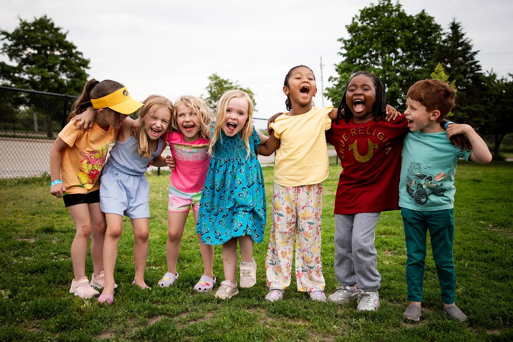 Group of seven children standing on grass with arms around each other, smiling and laughing outdoors.